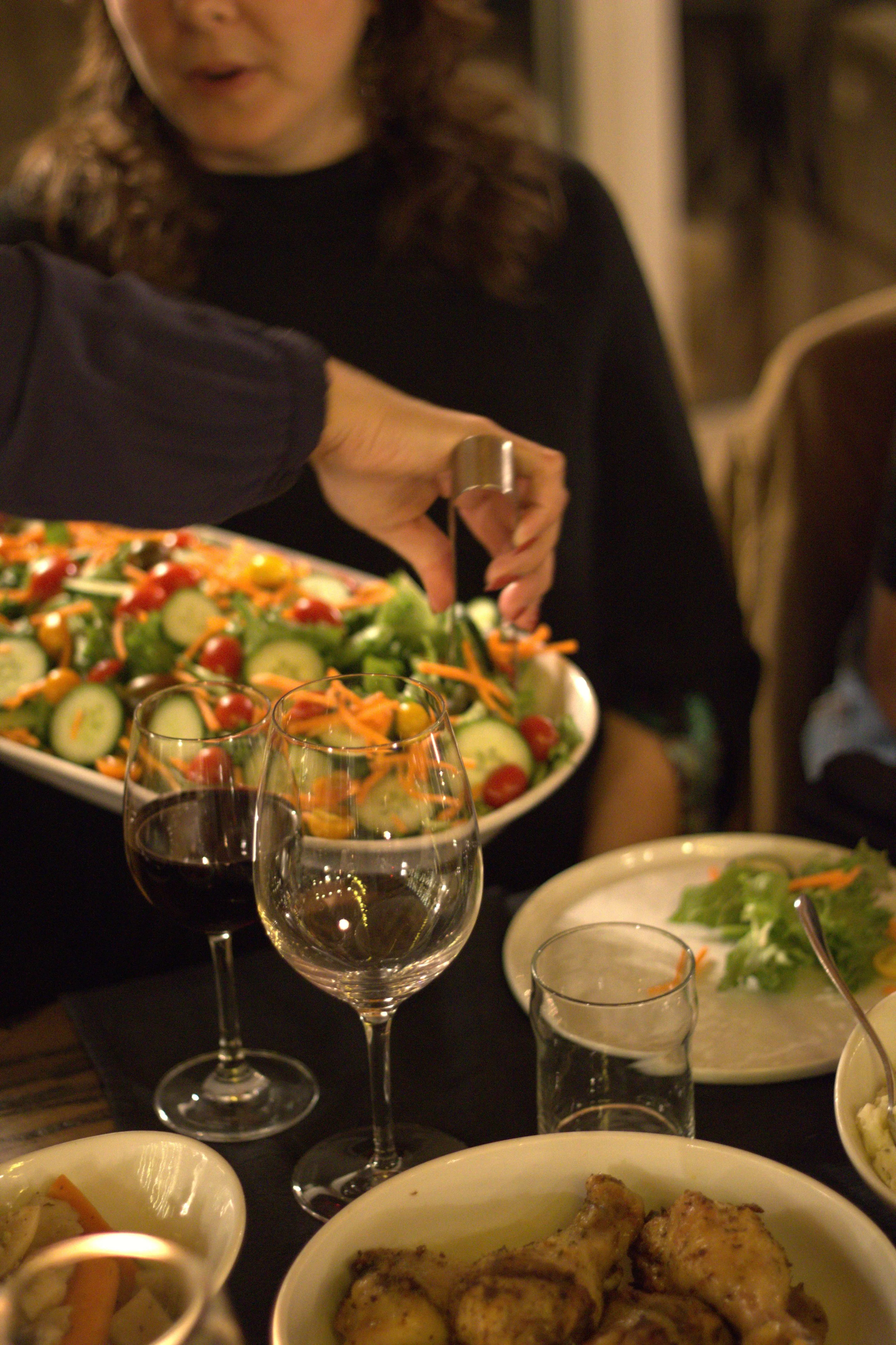 Person preparing a salad at a dinner table with various dishes and glasses of wine.