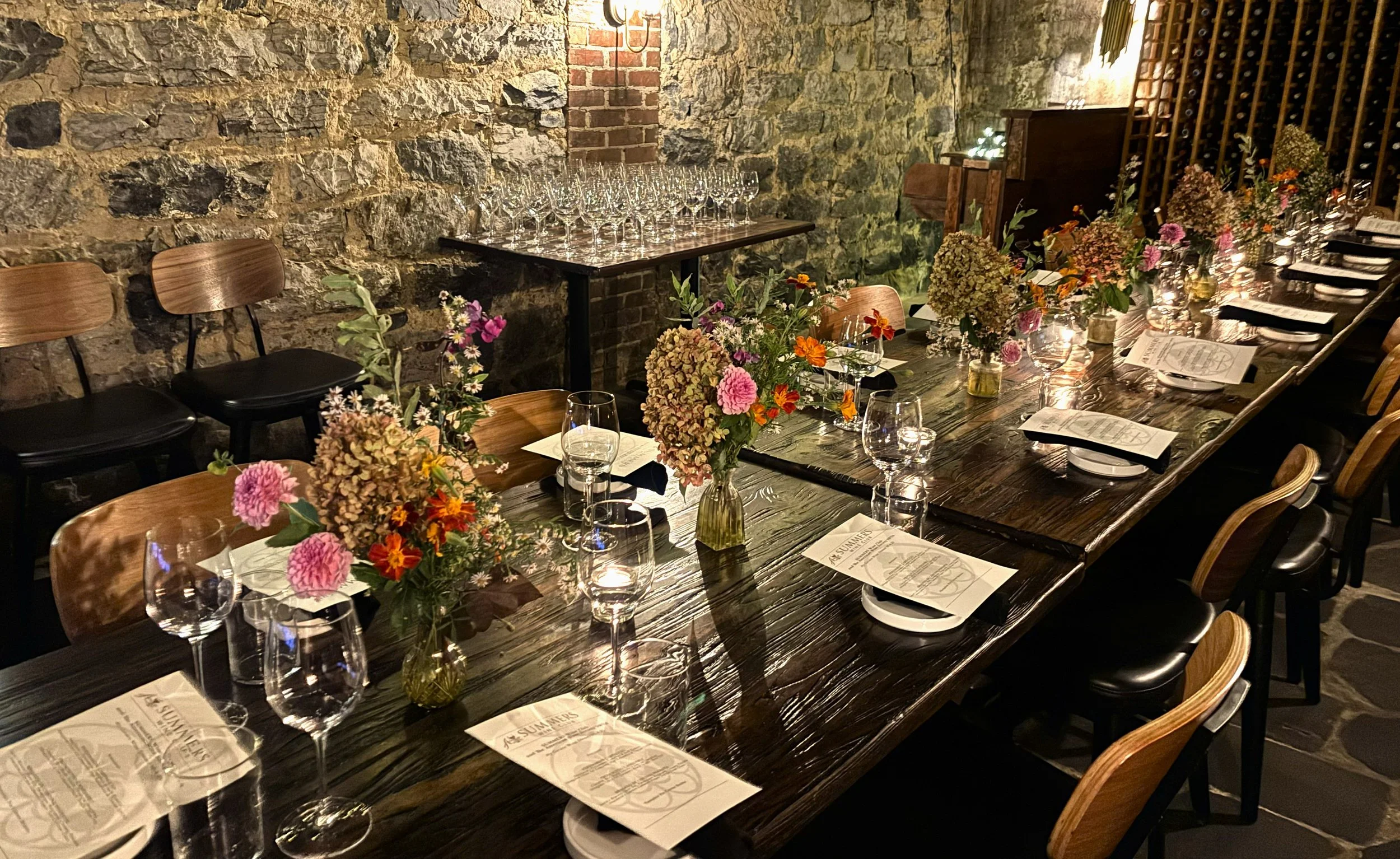 A long wooden dining table decorated with pink, orange, and purple flowers in glass vases, set with wine glasses, water glasses, plates, and menus, in a rustic restaurant with stone walls and a wine rack.