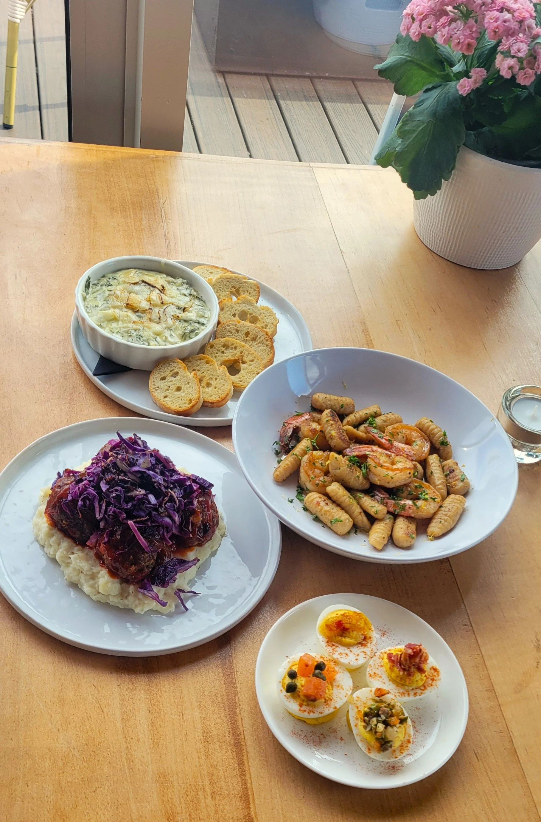 Assorted dinner dishes including a plate of  deviled eggs with toppings, a bowl of gnocchi with shrimp, mashed potatoes topped with meat and purple cabbage, a bowl of creamy spinach dip with bread slices, and a glass of water.