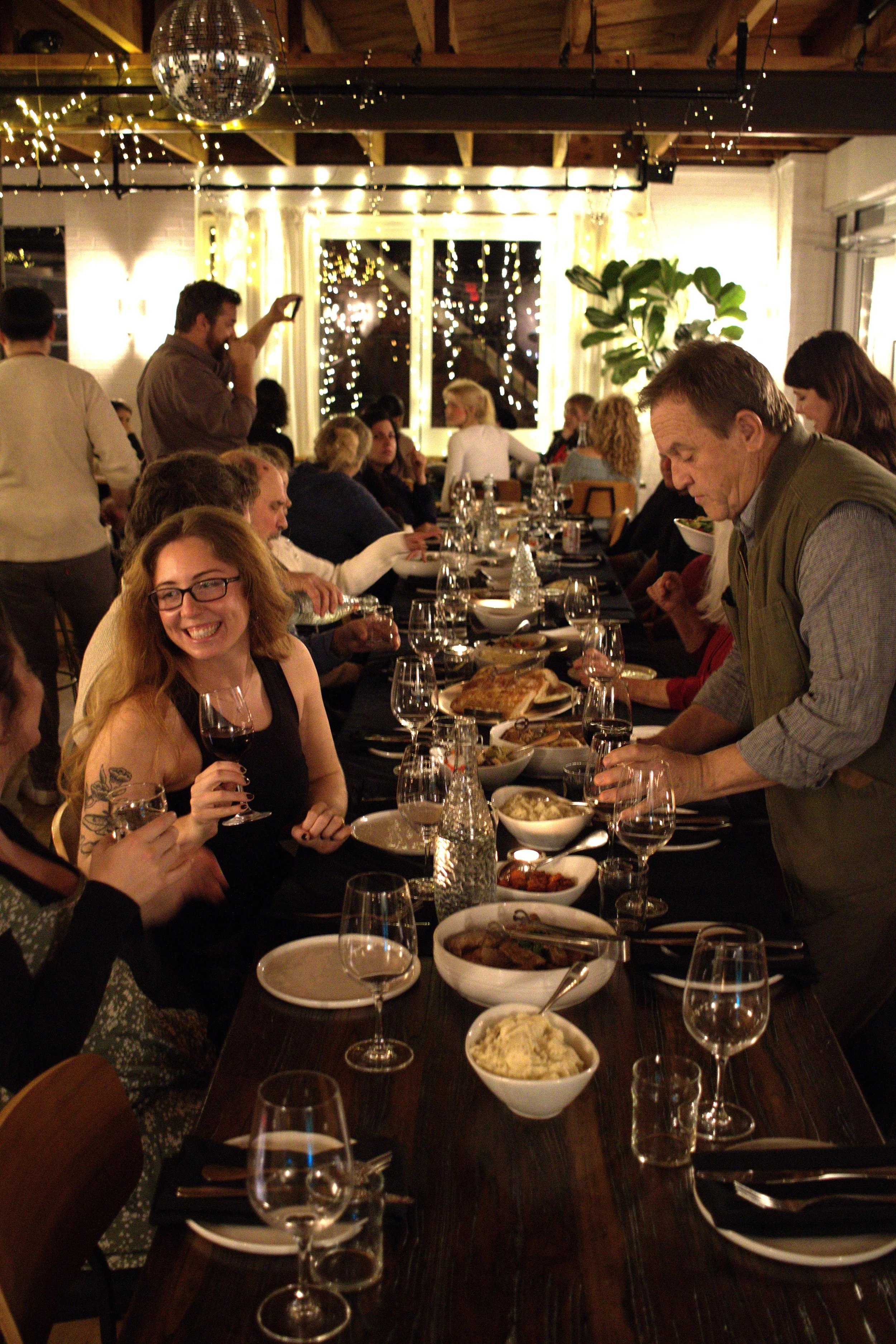 People enjoying a dinner party in a warmly lit room with string lights and a disco ball, seated at a long table with food and drinks.