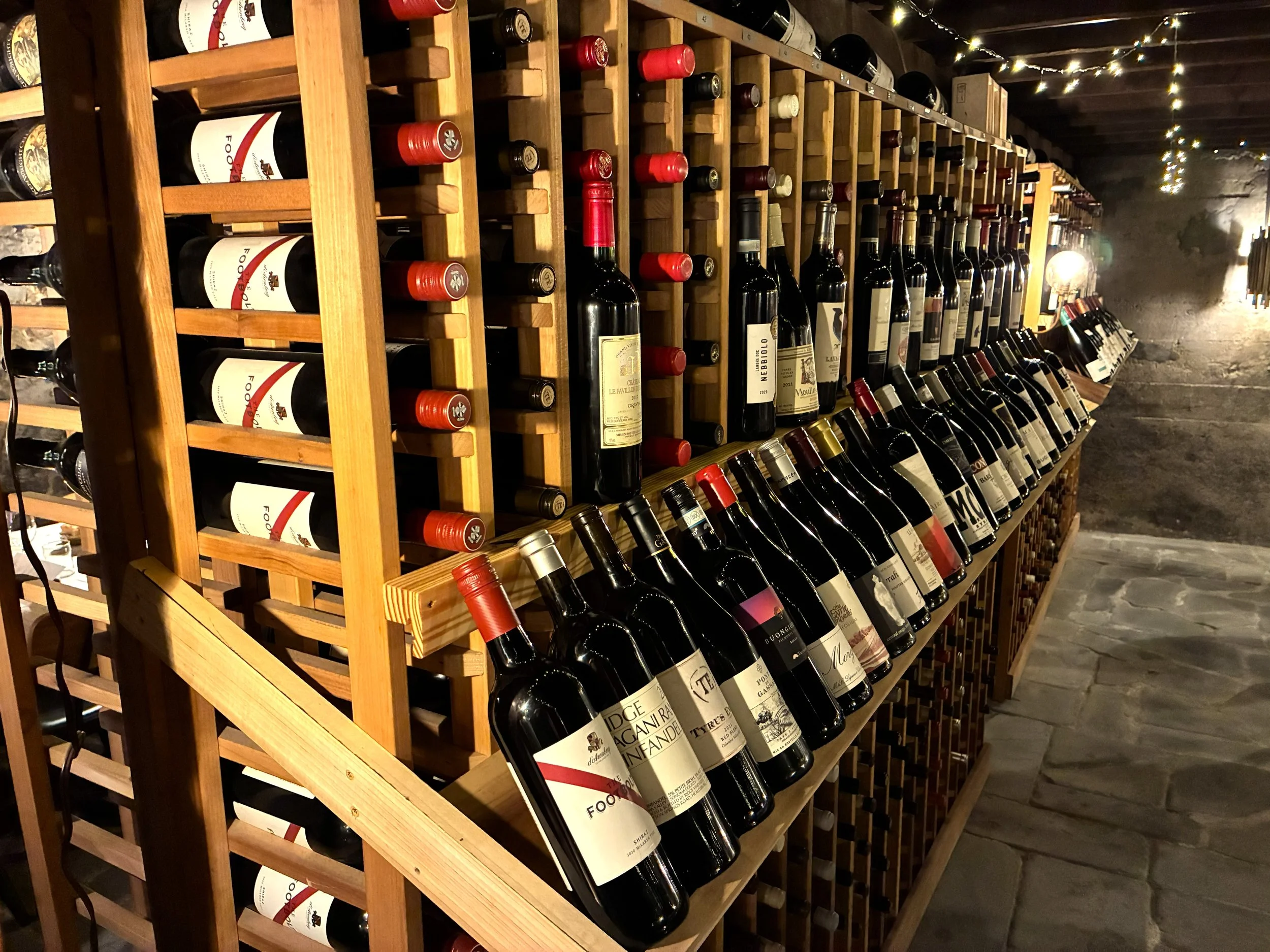 Wooden wine rack filled with red and white wine bottles, with some bottles laying horizontally and others vertically, inside a dimly lit cellar with string lights on the ceiling.
