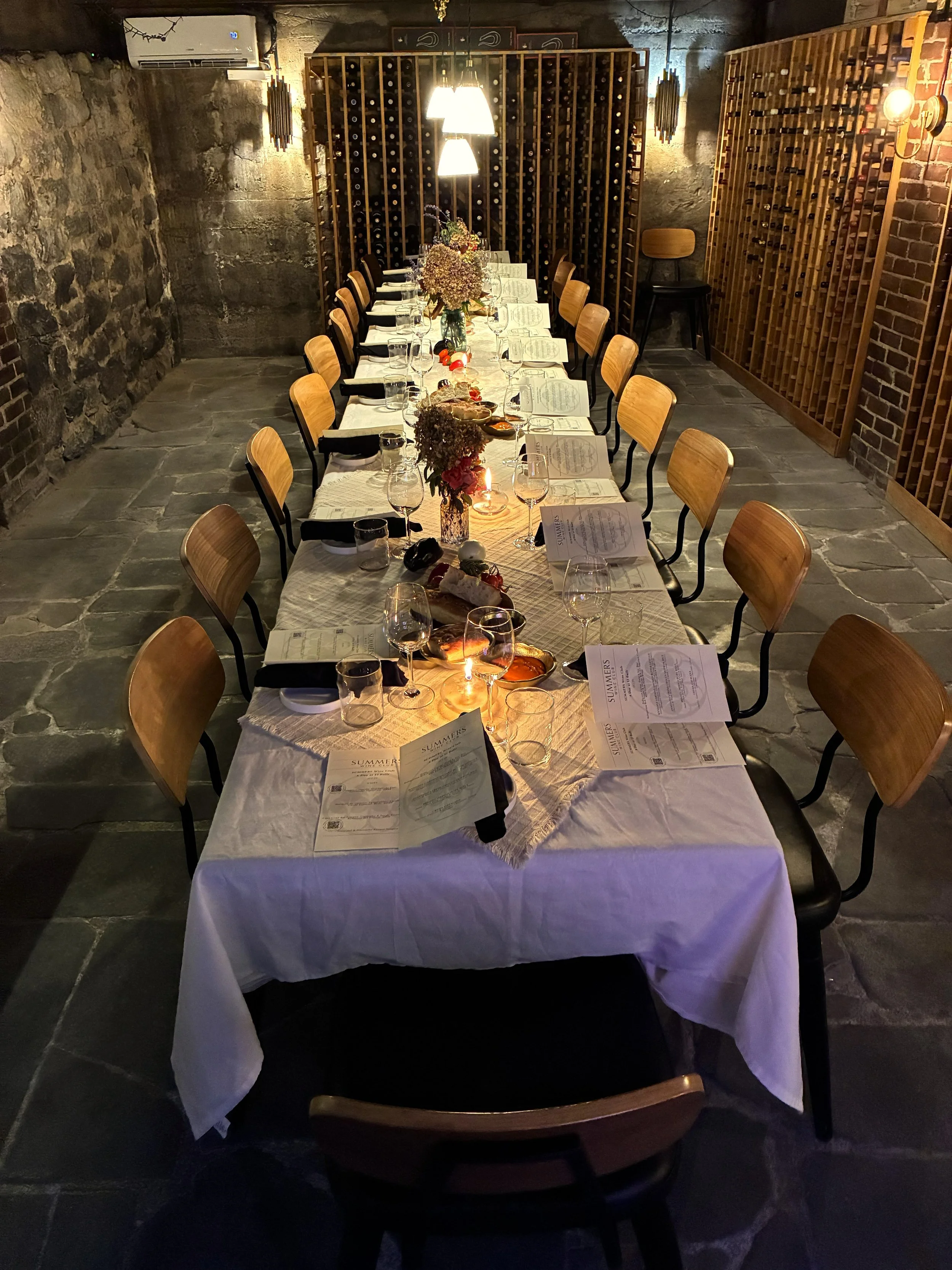 A cozy restaurant with exposed stone walls, lit by candlelight and pendant lights. A bartender is preparing drinks behind the bar, which is stocked with various bottles of alcohol. Empty tables with glasses, plates, and utensils are set for diners.