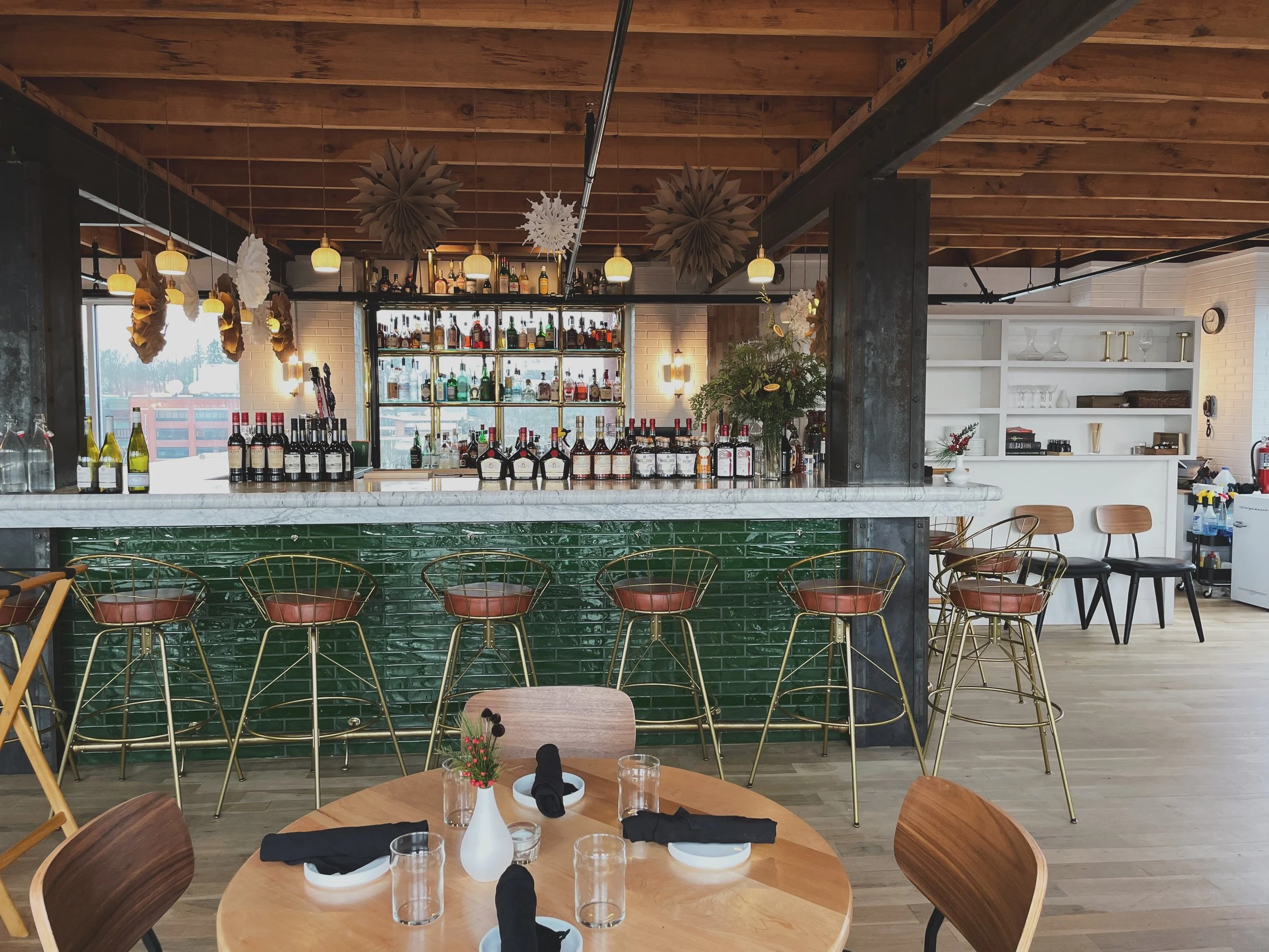 A modern restaurant interior with a bar area featuring gold chairs and a white marble countertop, adorned with bottles of alcohol. The dining area has round wooden tables set with black napkins, glasses, and small vases with flowers. The ceiling is wooden with hanging paper decorations, and there are large windows on the left side illuminating the space.