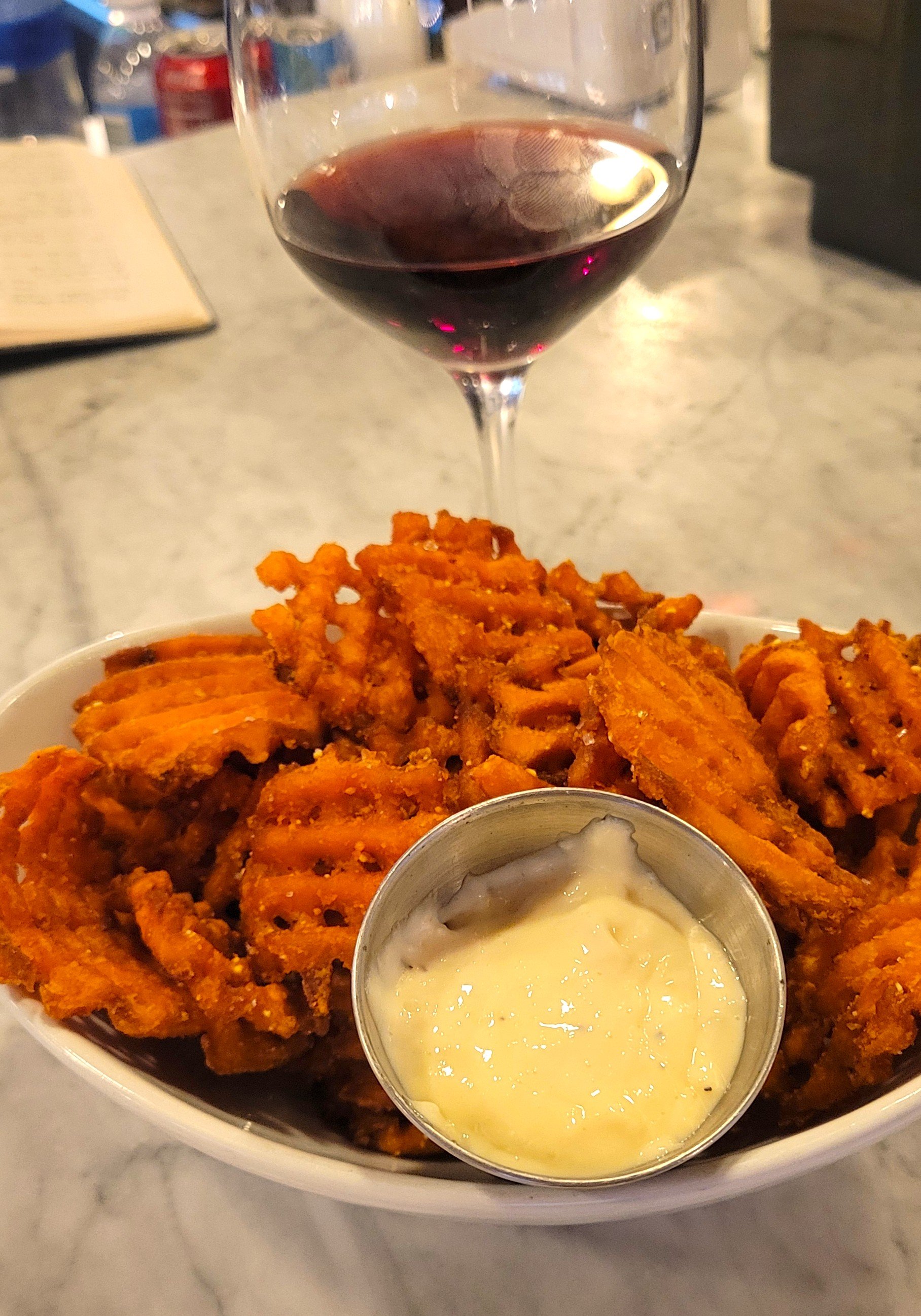 A bowl of waffle fries with a side of creamy dipping sauce, a glass of red wine, and a blurry background of a table with drinks and a menu.