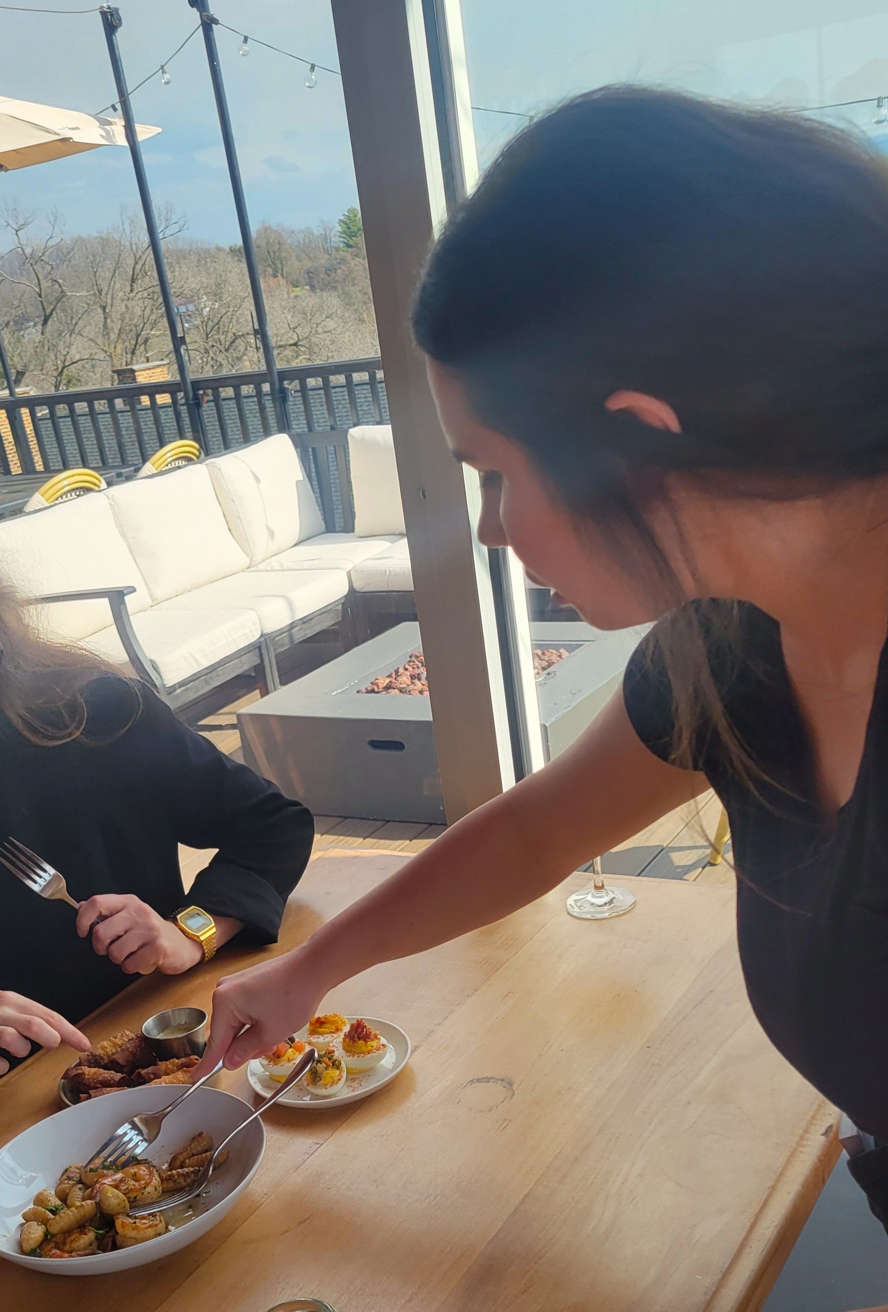 A woman serving food at a dining table with another person. Outside patio with white cushioned seating and string lights visible through large glass windows.