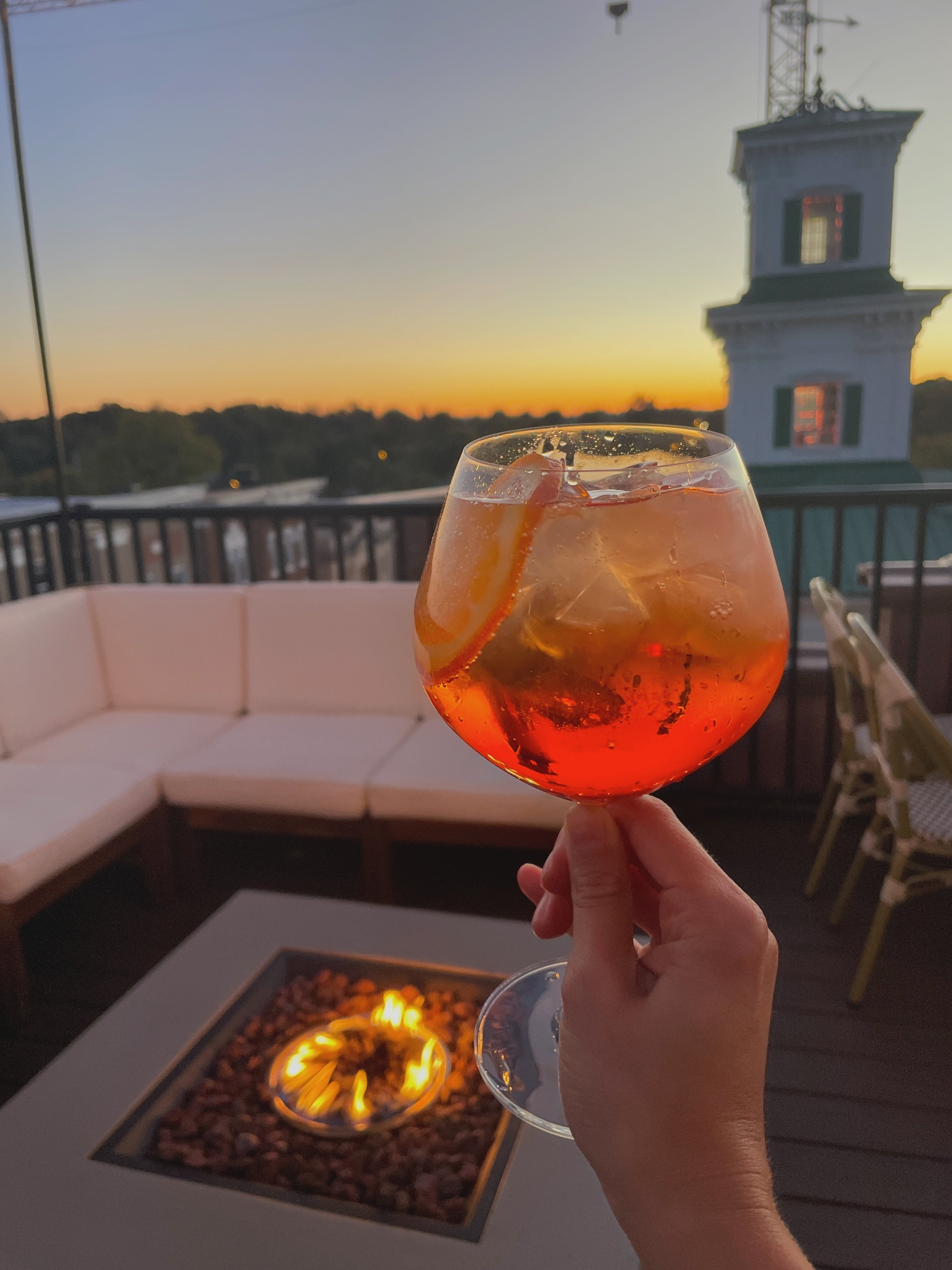 A person holding a glass of Aperol spritz with ice and orange slices on an outdoor patio at sunset, with a round fire pit, white courtyard furniture, and a clock tower in the background.
