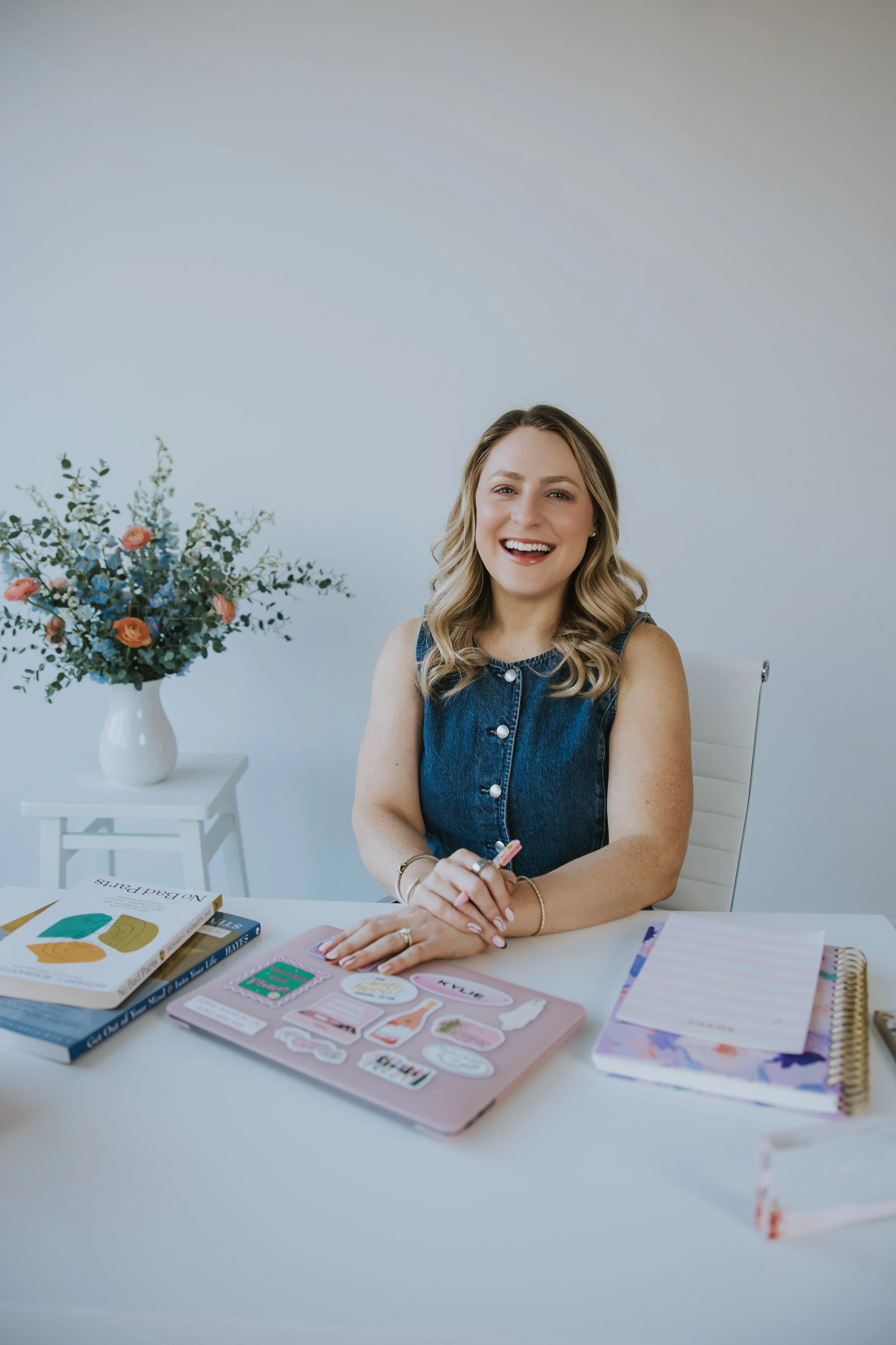 Kylie Lambert, therapist, smiling and sitting at a desk with a laptop, books, and a vase of flowers beside her.