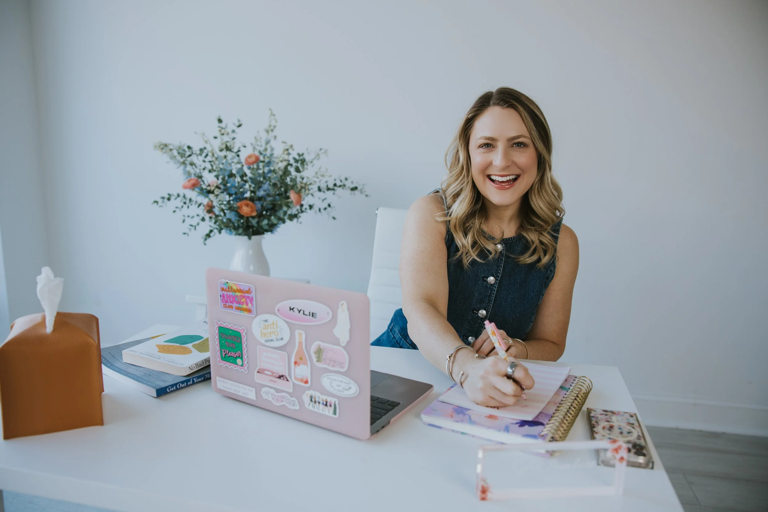 A woman with blonde hair smiling while sitting at a desk filled with books, notebooks, and laptop with stickers, in a room with a large flower arrangement in a white vase on the desk.