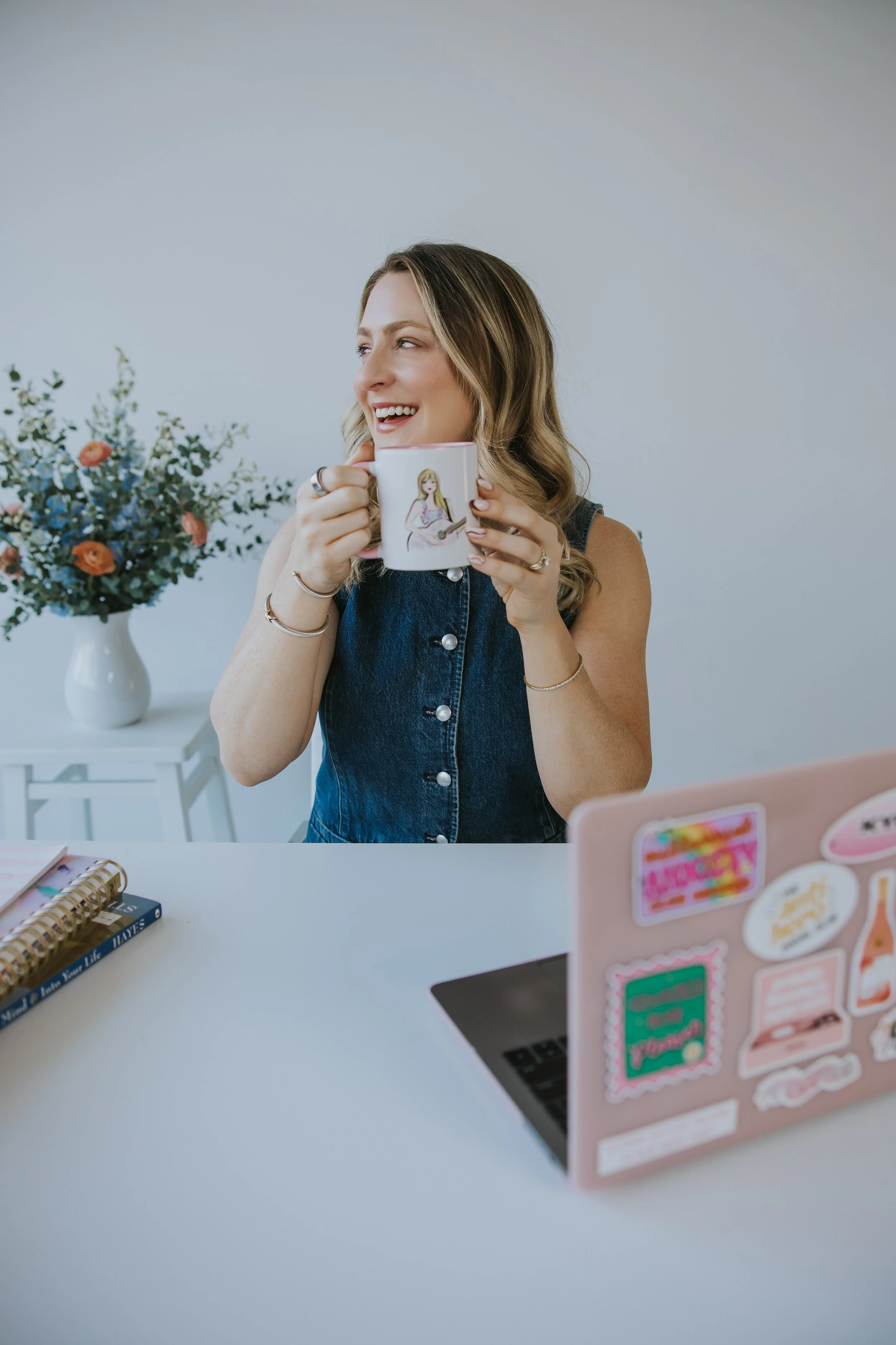 Kylie Lambert, therapist, laughing while holding a mug, sitting at a desk with flowers and a laptop nearby