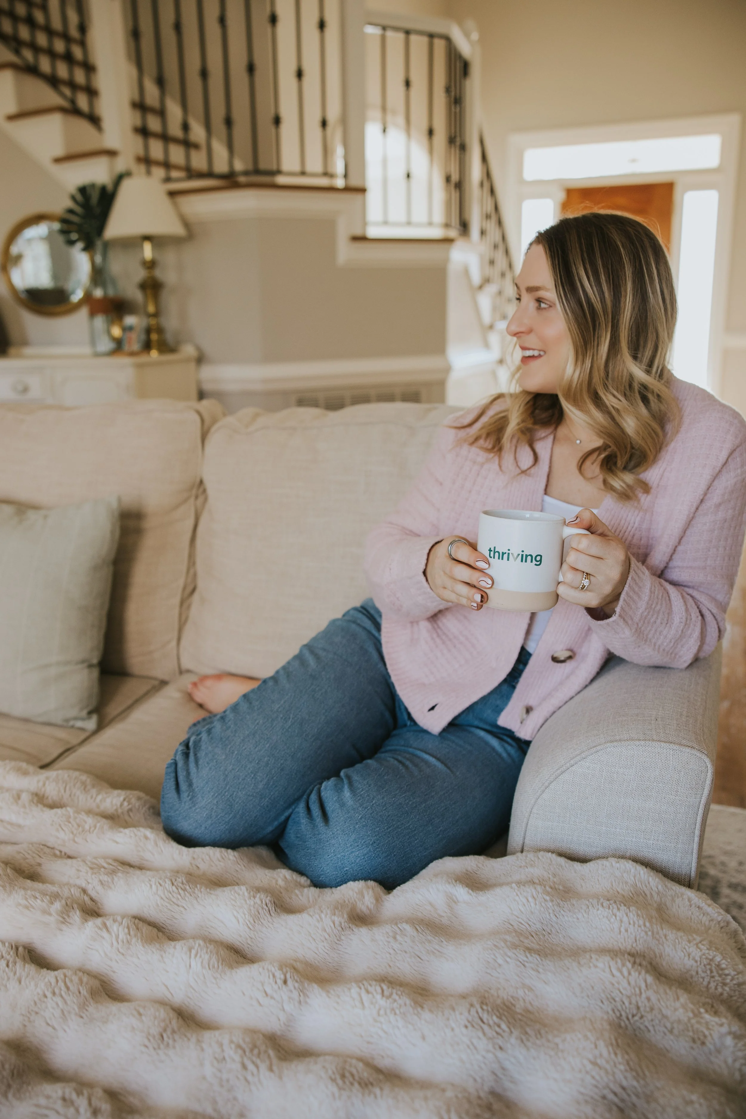 Kylie Lambert, therapist, sitting on a couch holding a mug that says 'thriving,' in a bright entryway with a staircase in the background.