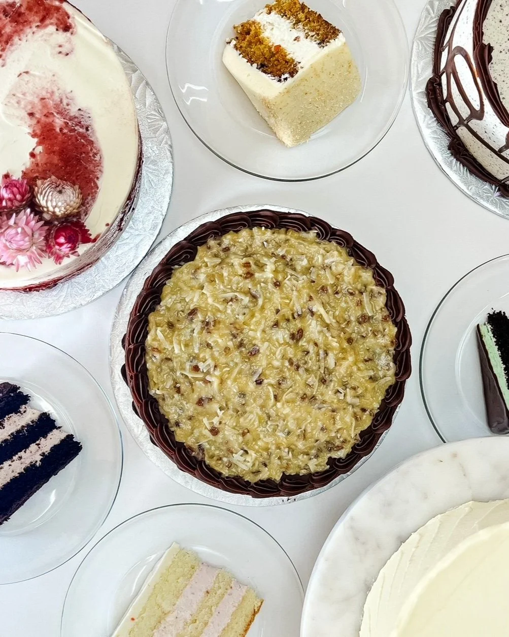 Assorted cakes on a table, including a fruit-topped cake, a white cake with chocolate drizzle, a layered chocolate cake, a white frosted cake, and a German chocolate cake with coconut pecan topping..