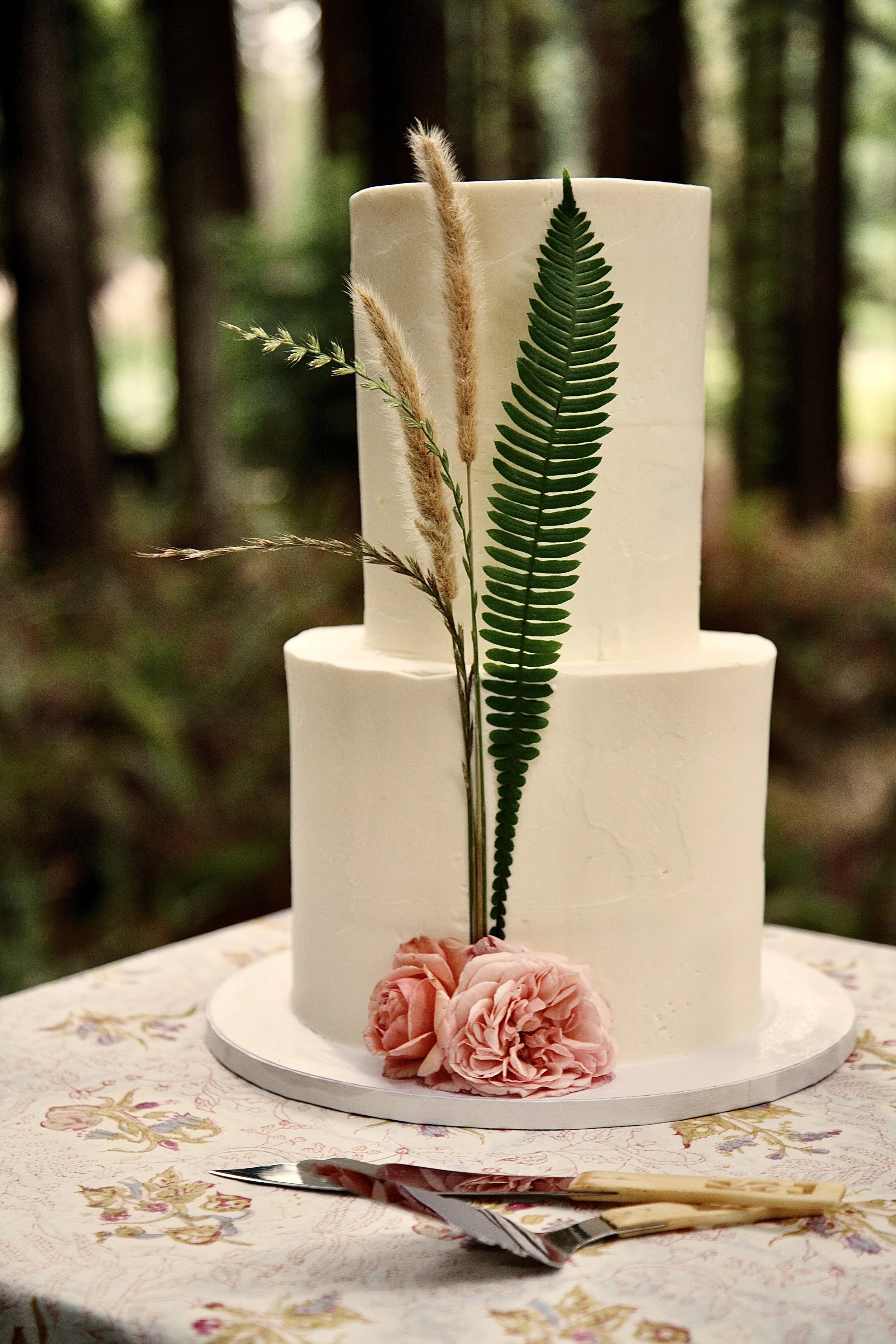 A two-tiered white cake with pink flowers, green fern leaves, and pampas grass