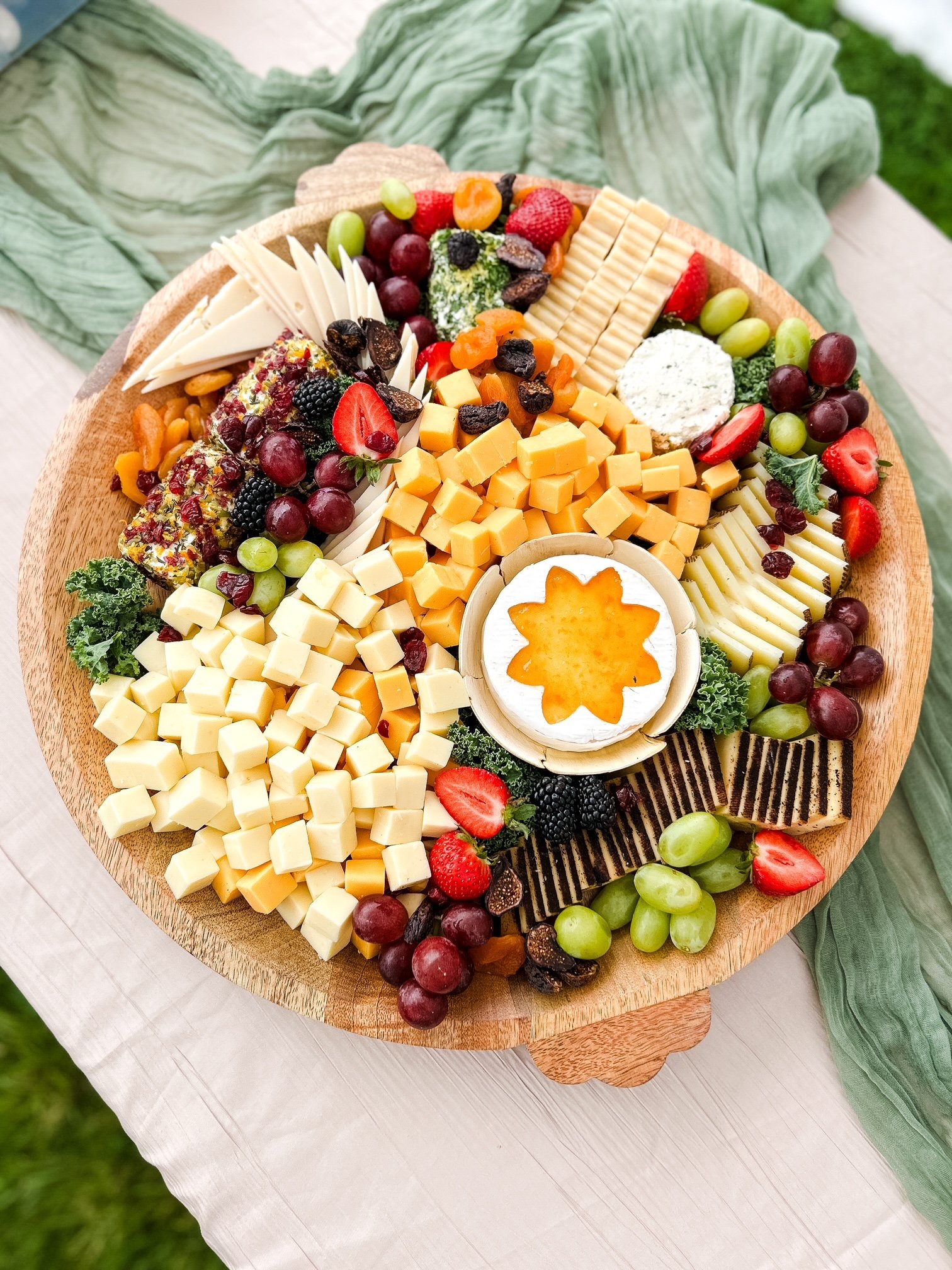 A cheese and fruit platter arranged on a round wooden tray with grapes, strawberries, blackberries, dried and fresh fruits, various cheeses, and crackers.