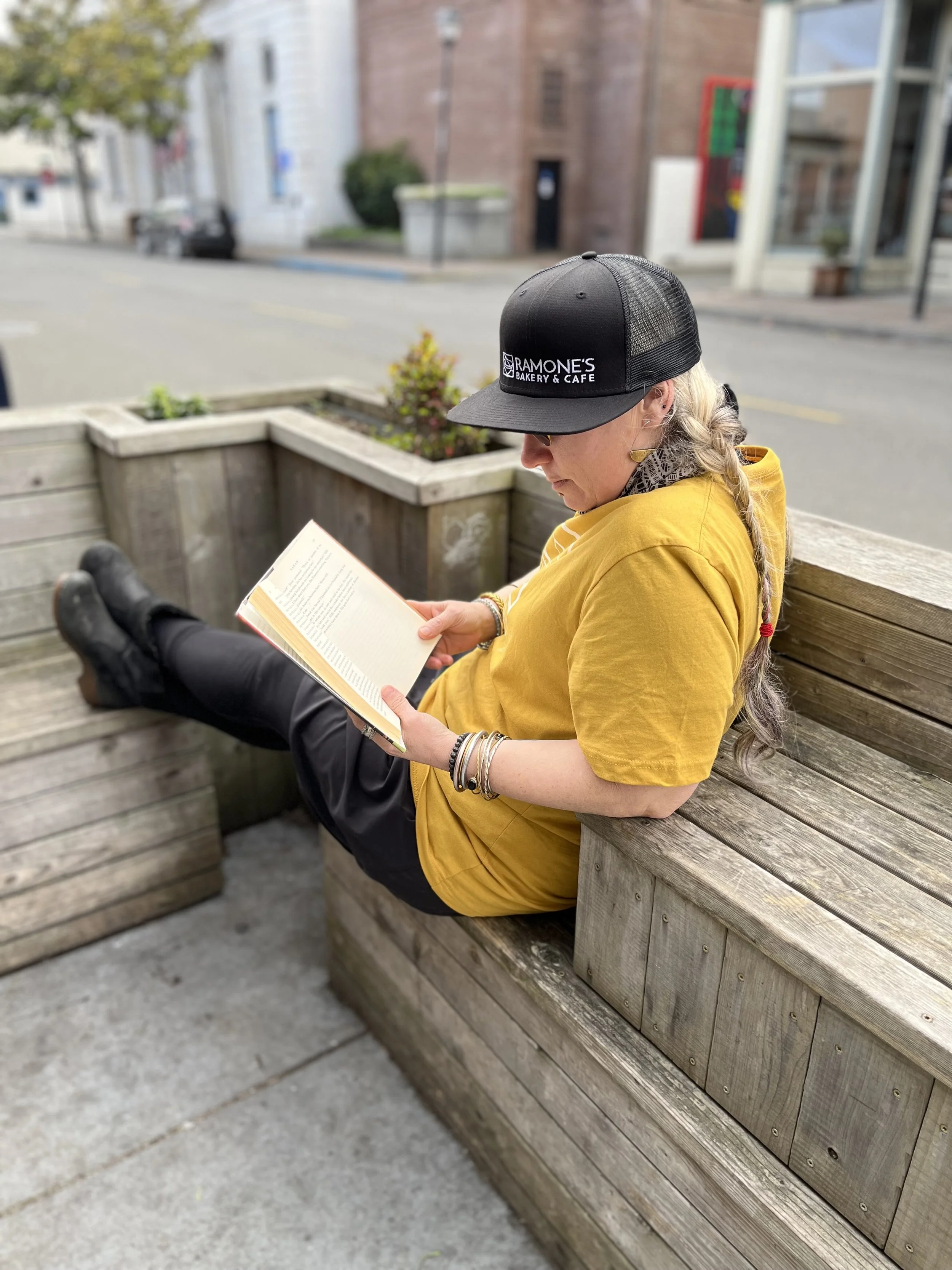 Person wearing black trucker hat, lounging on bench reading a book