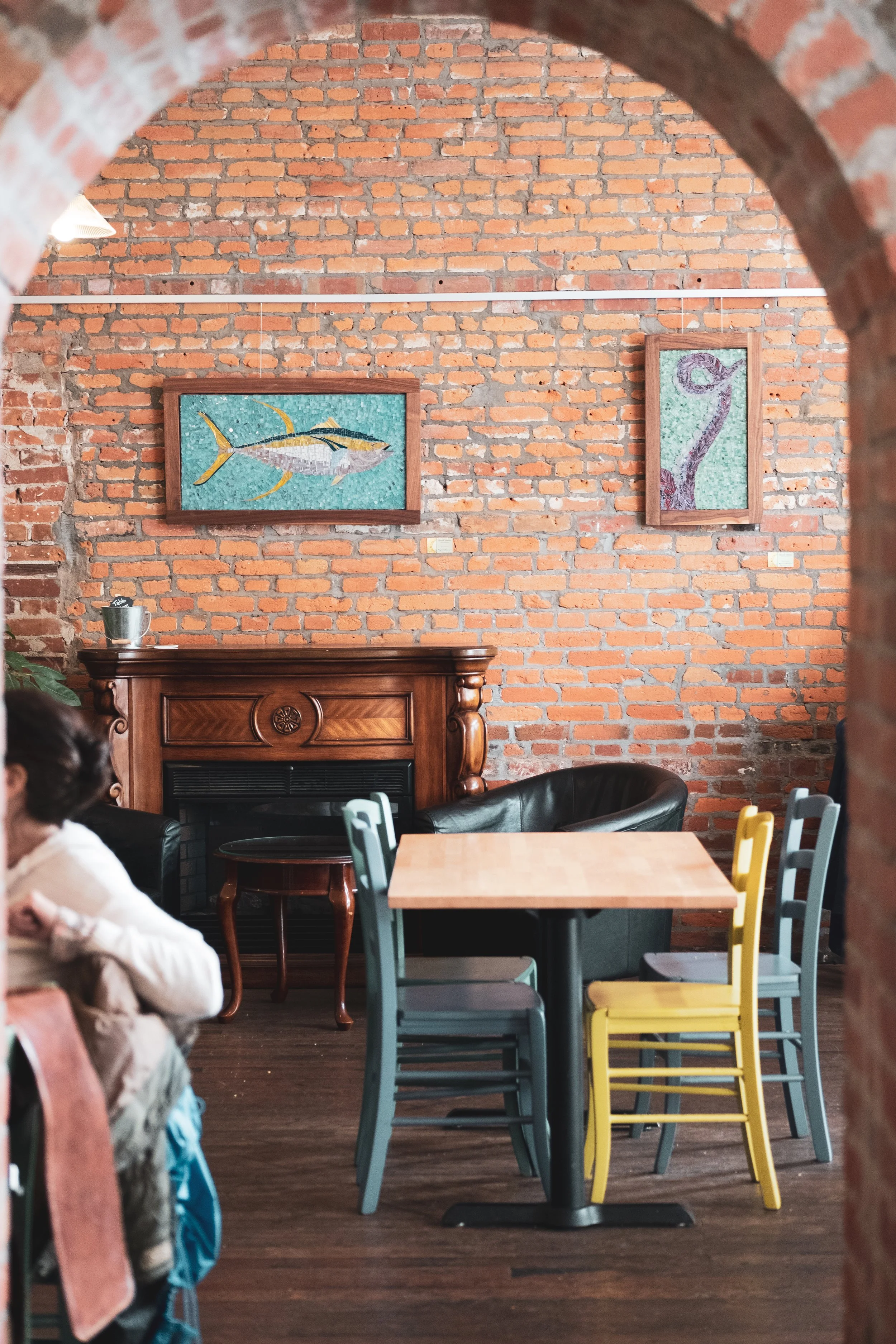 A cozy cafe interior with a brick wall, colorful chairs around a wooden table, artwork of a fish and an abstract design on the wall, and a fireplace with a decorative wooden mantle.
