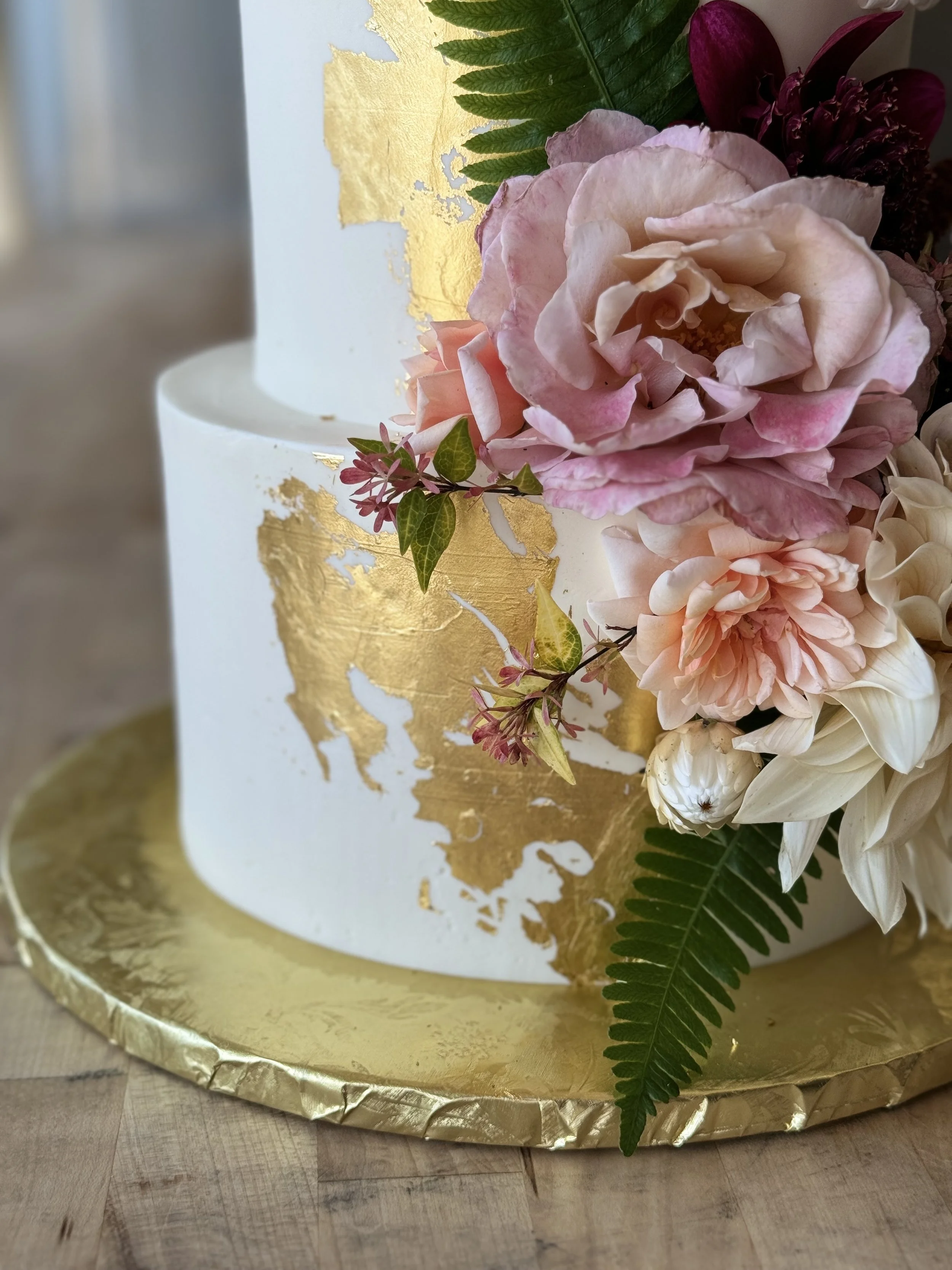 A two-tiered white cake decorated with gold leaf accents and fresh flowers