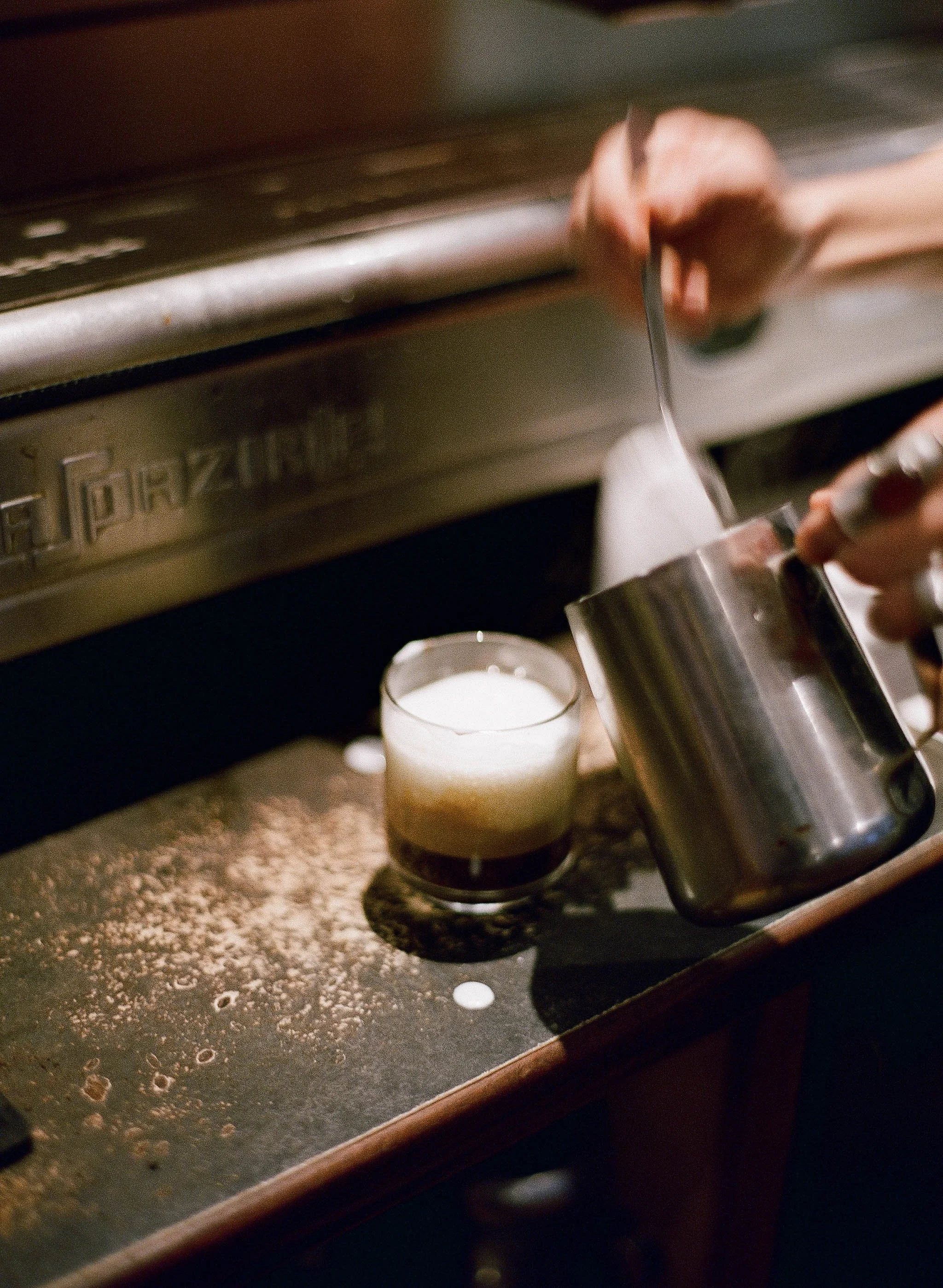 A person pouring steamed milk into a glass of espresso coffee, creating a layered drink with foam on top, on a barista's work surface.