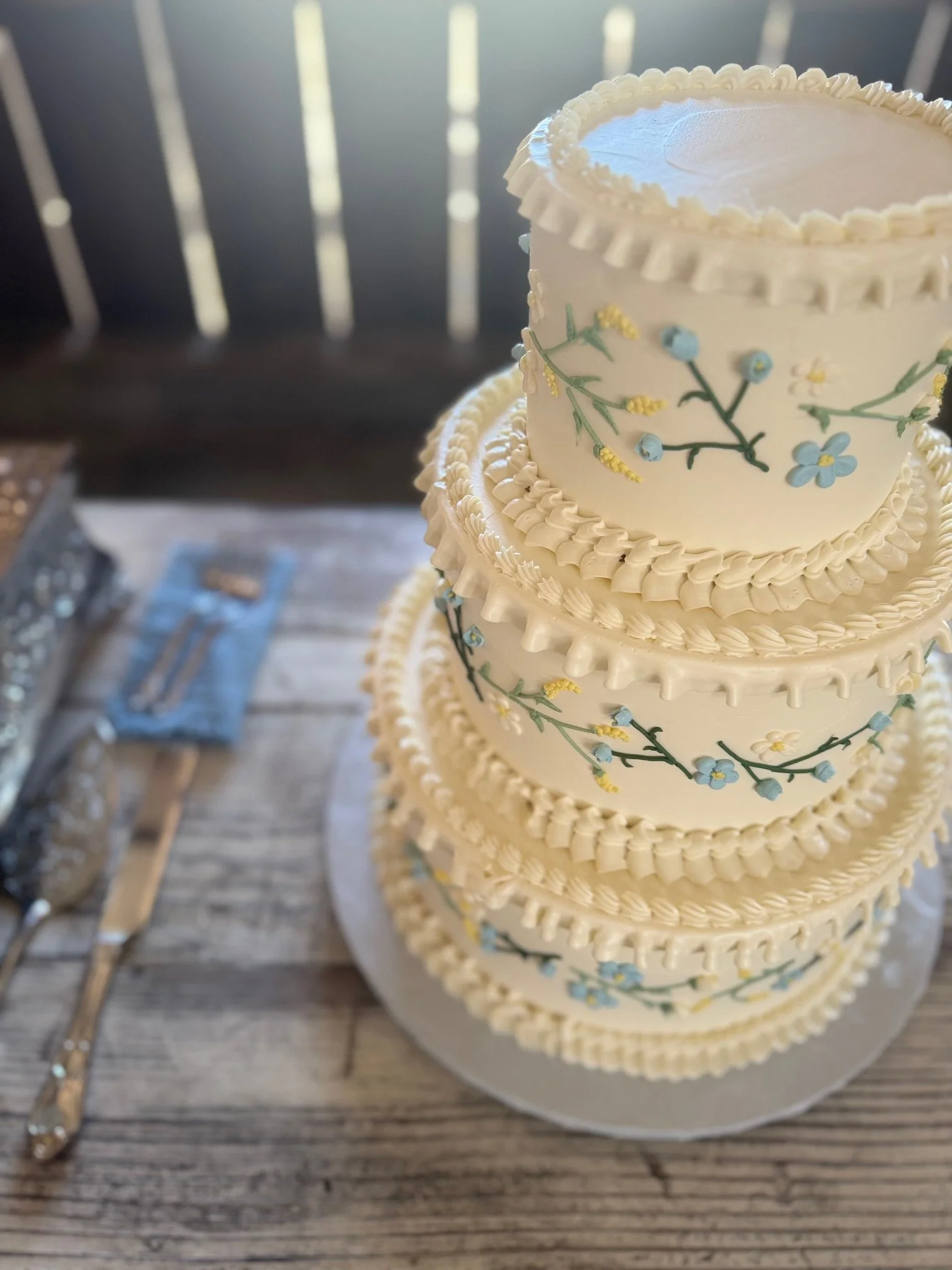Four-tiered wedding cake decorated with white icing and delicate floral patterns with blue and yellow flowers and green vines.