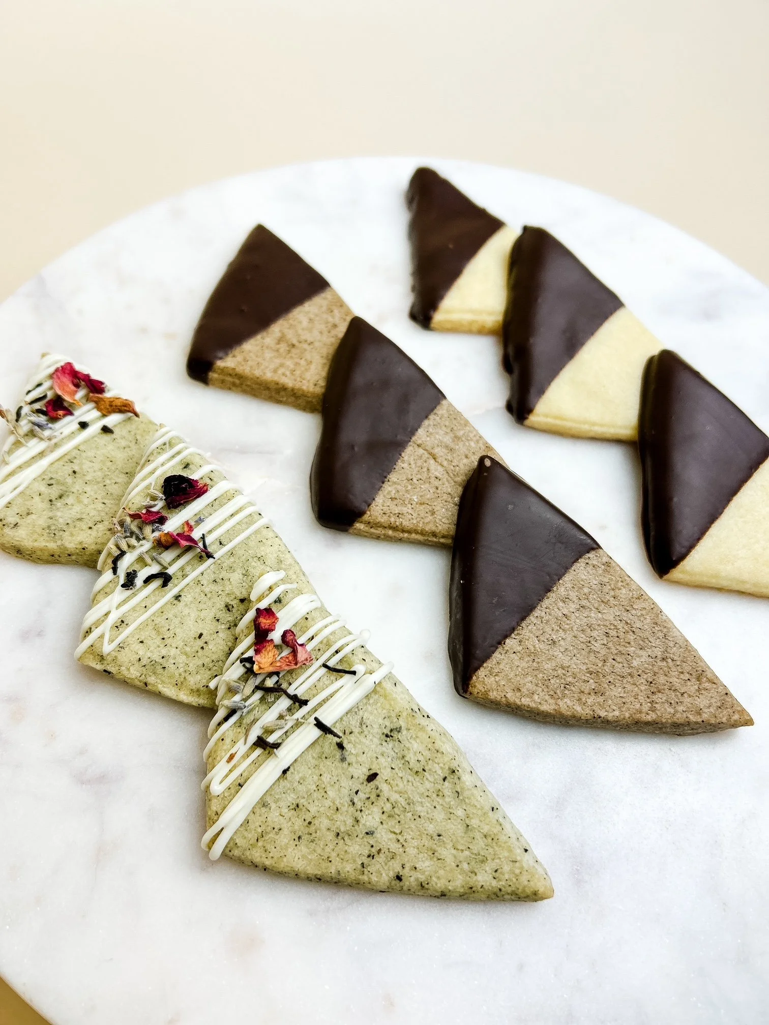 Three types of triangle-shaped shortbread cookies on a white marble platter. Earl gray shortbread cookies drizzled with white chocolate and sprinkled with edible flowers, espresso shortbread and plain shortbread dipped in chocolate.