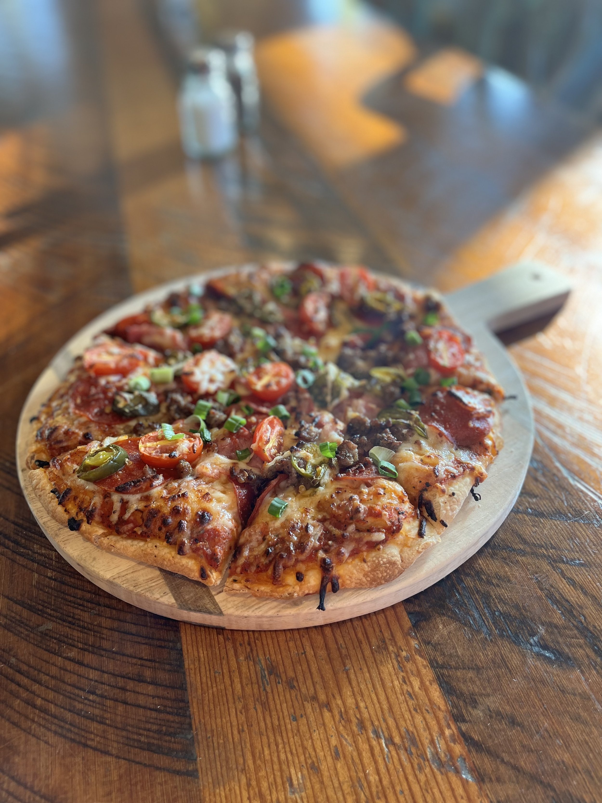A pizza with various toppings including cherry tomatoes, green onions, and sliced green peppers on a round wooden serving board on a rustic wooden table.
