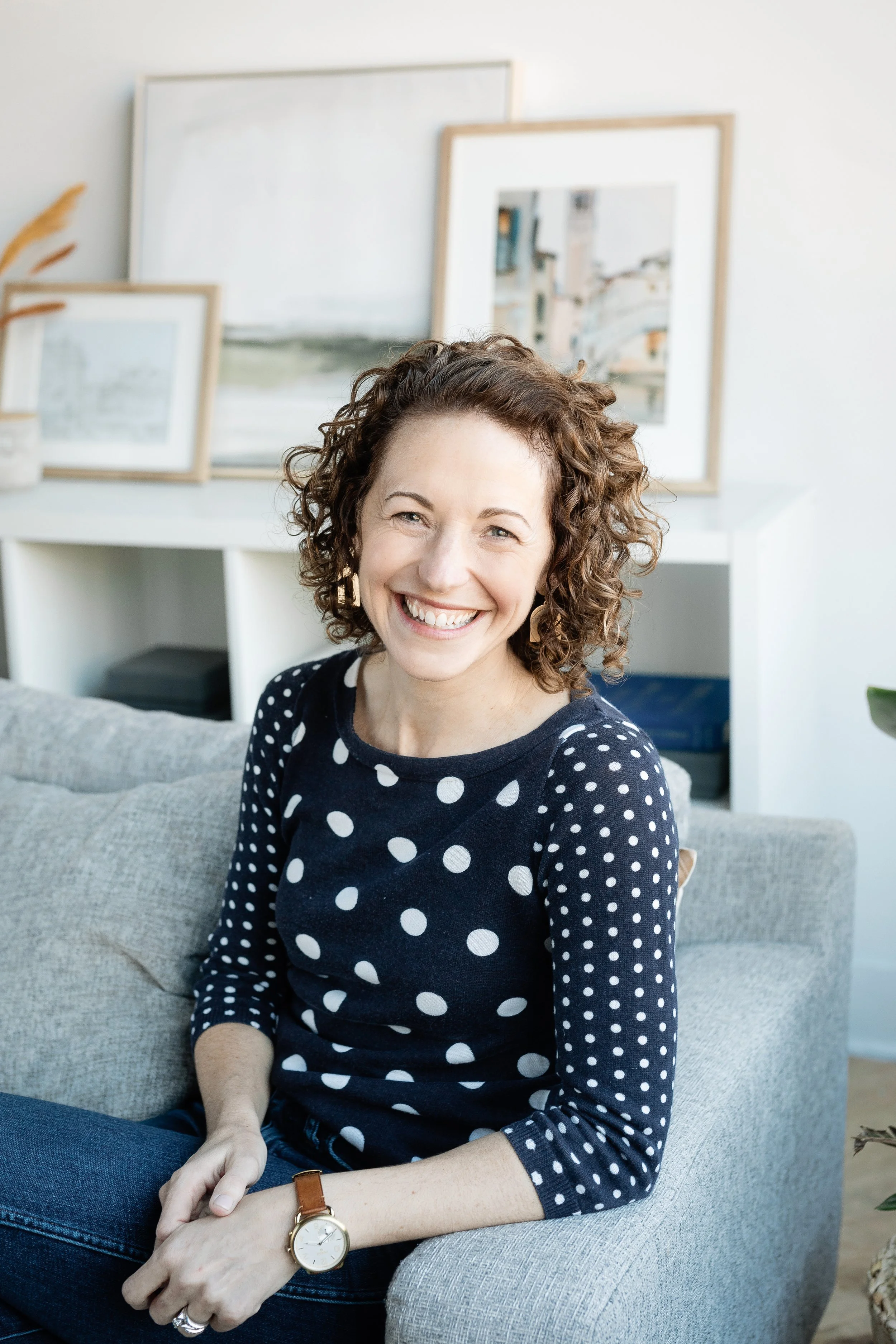 A woman with curly hair smiling, sitting on a gray couch in a room with framed pictures on the wall behind her.