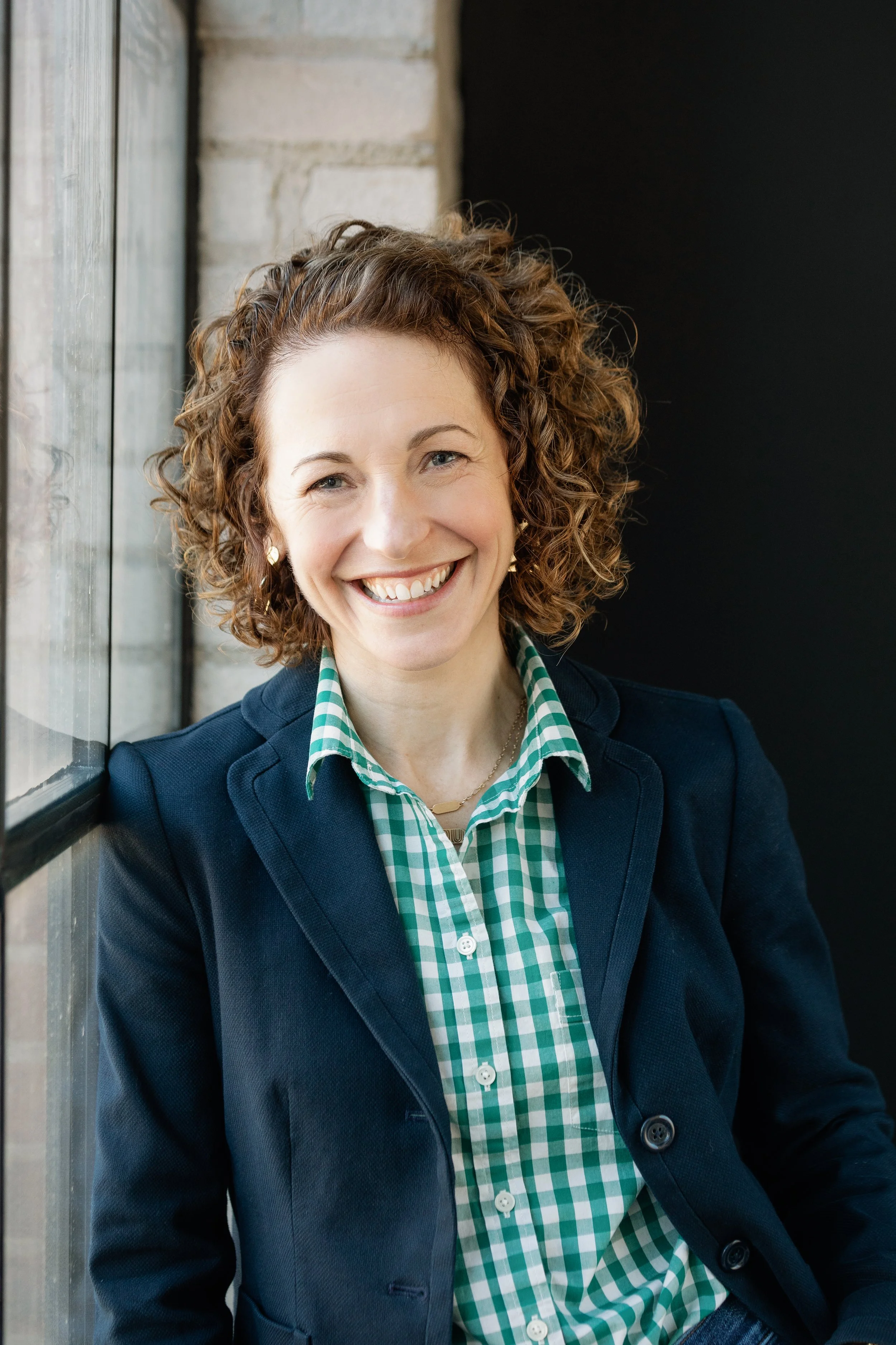 A woman with curly reddish hair smiling, wearing a green and white checkered shirt and a navy blazer, standing next to a window with a brick wall in the background.