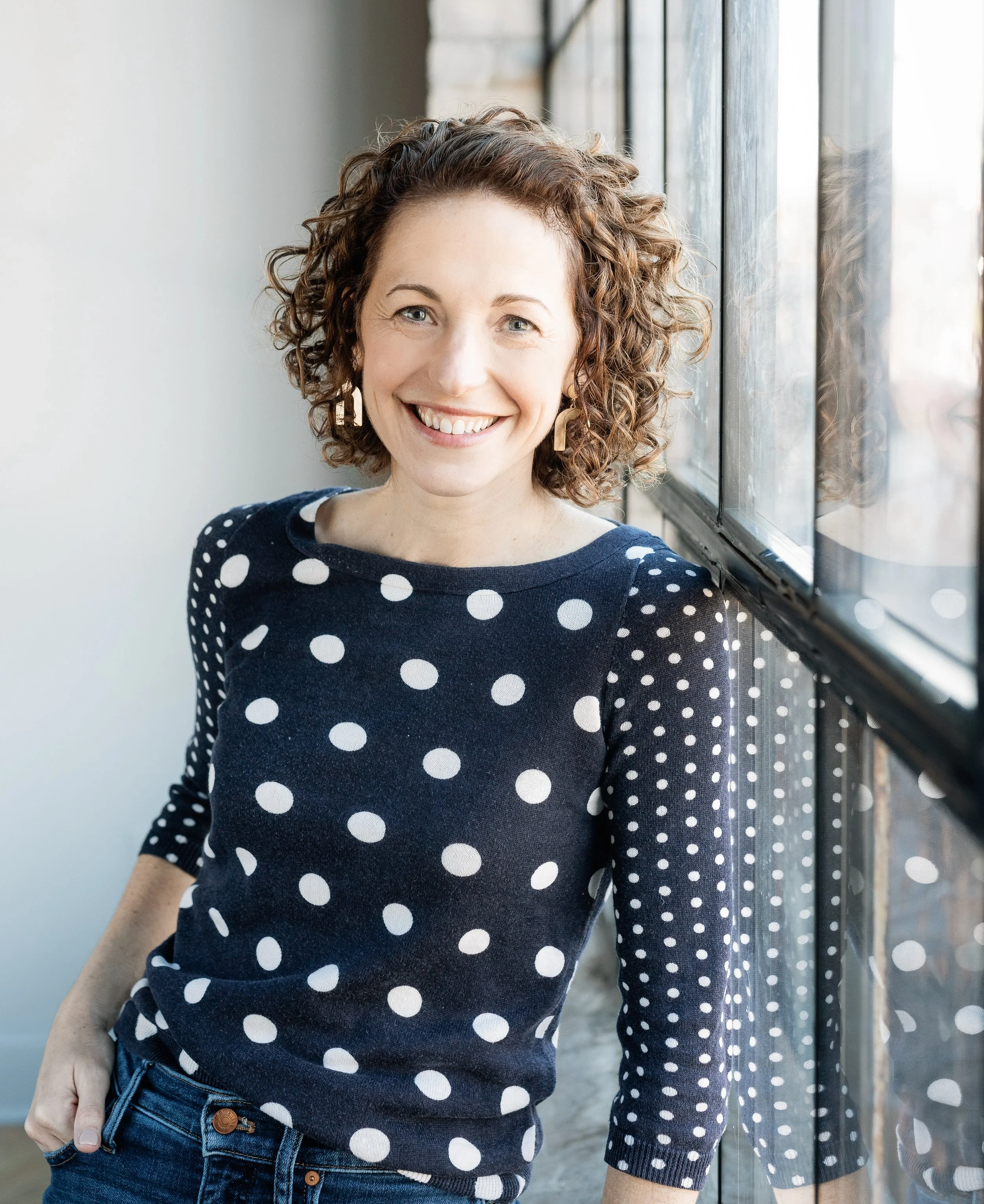 A woman with curly hair smiling, standing by a window with a brick wall outside, wearing a navy blue sweater with white polka dots and blue jeans.