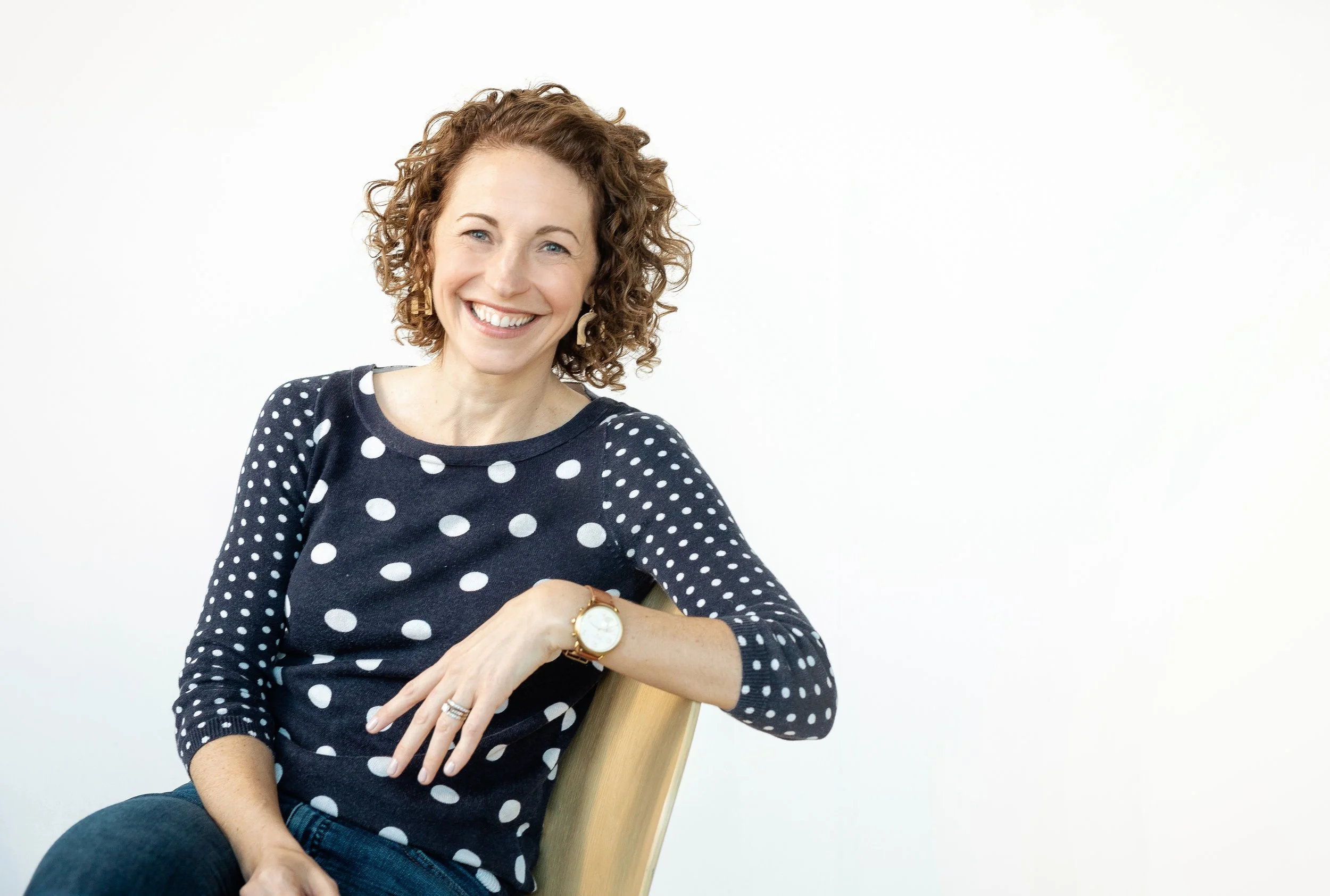 A woman with curly brown hair, smiling, wearing a navy blue sweater with white polka dots, sitting on a wooden chair against a white background.