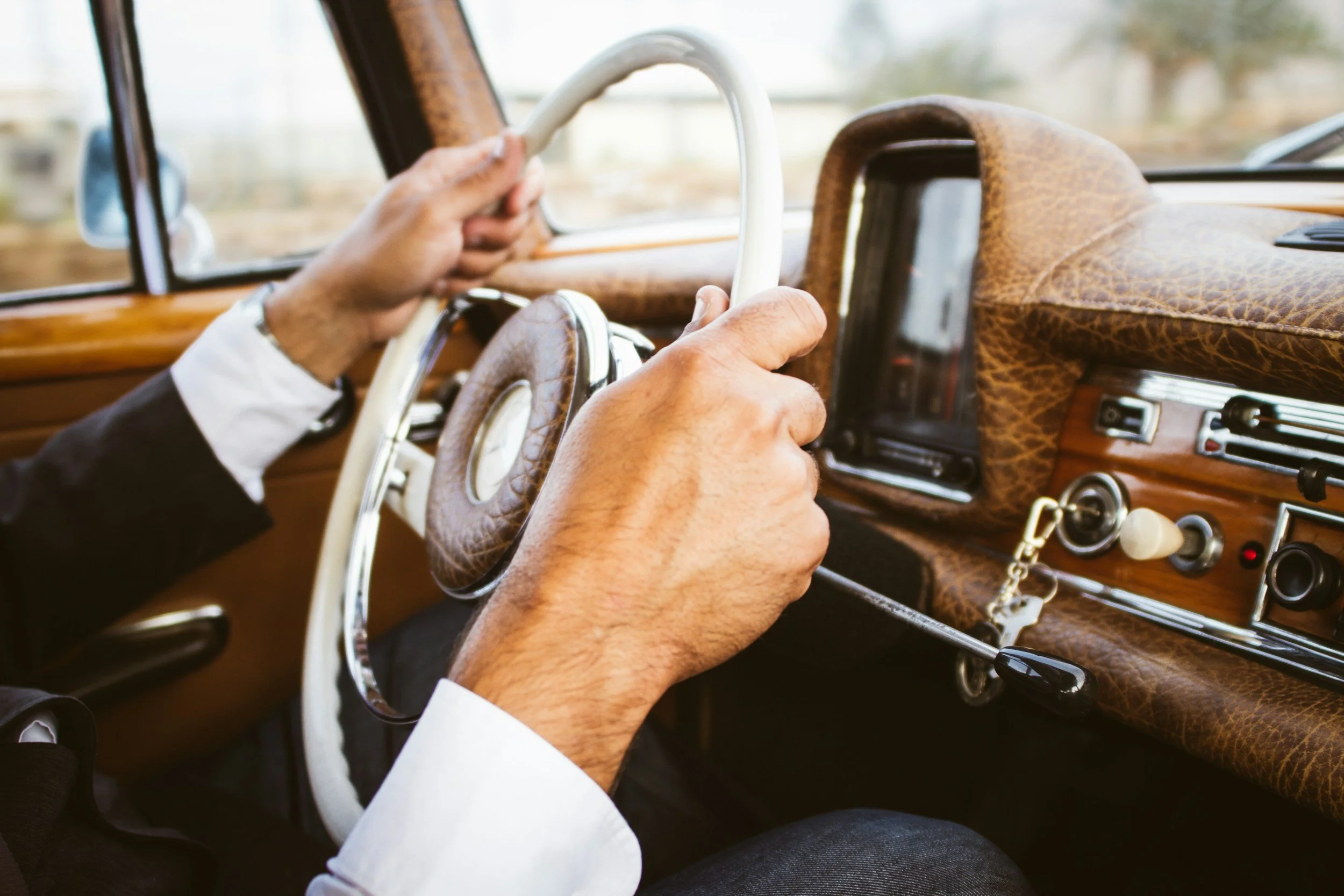 A person with light skin, wearing a white shirt and a dark jacket, is driving a vintage car with a wooden dashboard and steering wheel. The driver is holding the white steering wheel with both hands. The car has a classic look with brown leather and wood accents on the dashboard and headliner. The outside scenery is blurred through the windshield and side window.