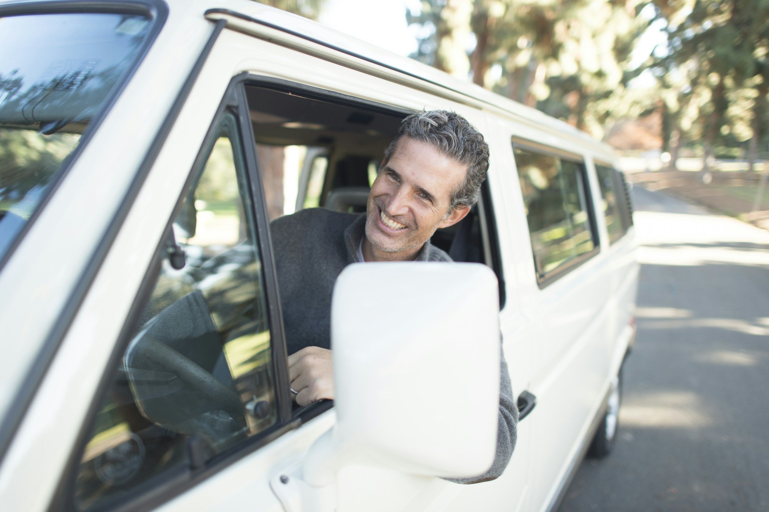 A man smiling and leaning out of the driver's side window of a white vehicle parked outdoors with trees in the background.