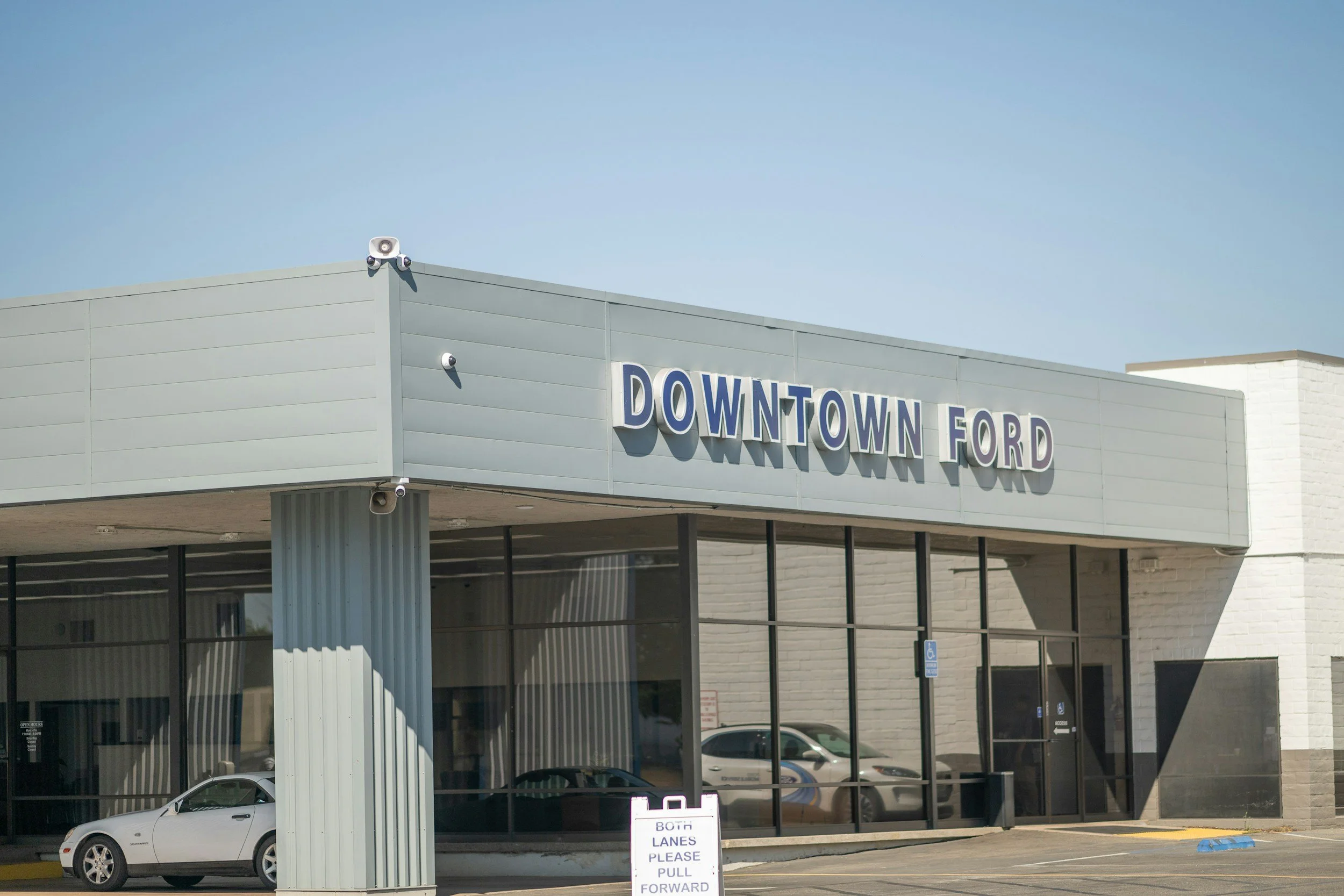 A modern car dealership building with large glass windows and a sign that reads "DOWNTOWN FORD." There is a white sports car parked in front of the building, and a sign near the entrance indicates to pull forward. The sky above is clear and blue.