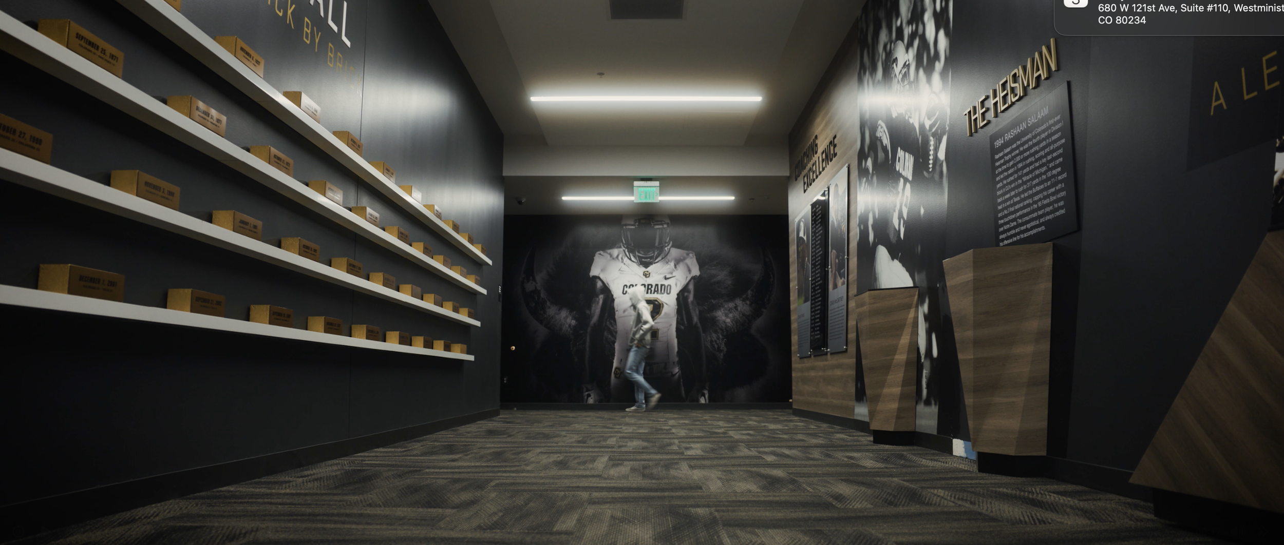 An indoor hallway with black walls and wood flooring, featuring a large mural of a football player in a Colorado Buffaloes uniform. Shelves with yellow boxes are on the left wall, and informational displays are on the right. An emergency exit sign is