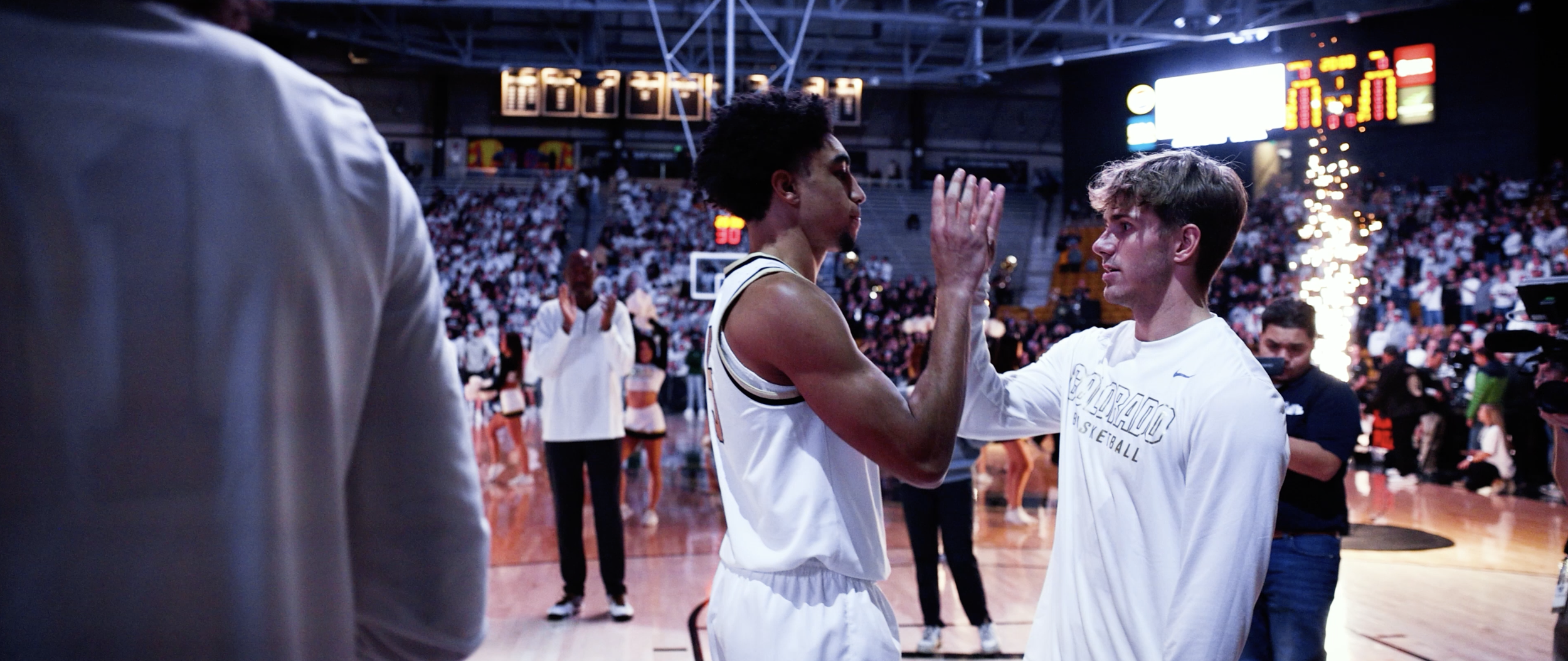 Two basketball players in white jerseys giving high fives on a basketball court, with a crowd in the background and scoreboard overhead.