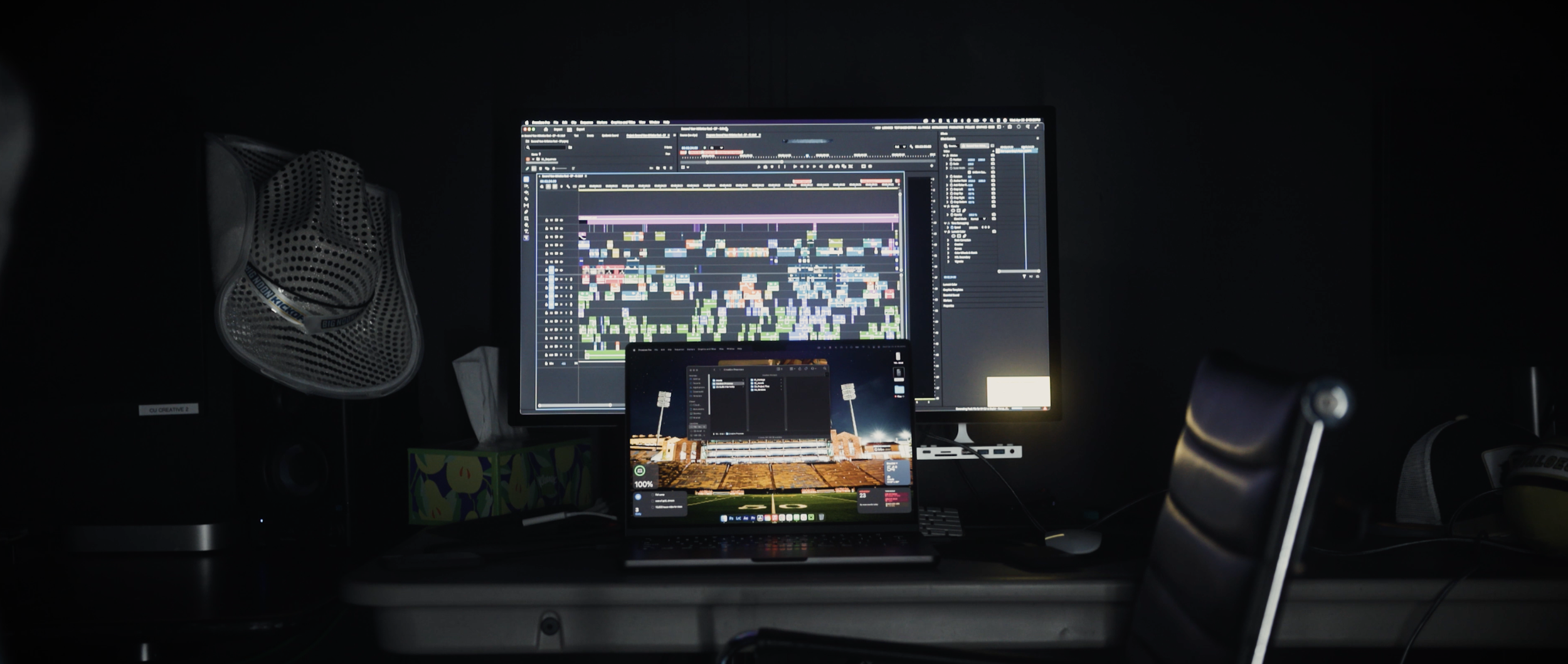 Computer setup with a large monitor displaying a music production software, a smaller laptop showing a stadium background, a speaker, a fan, and other miscellaneous items in a dark room.