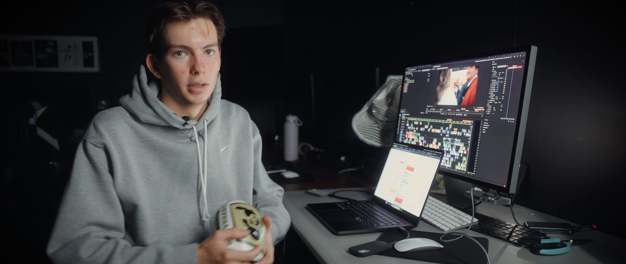 A young man in a gray hoodie sitting at a desk with dual monitors, a laptop, and editing software, holding a white container with black and yellow design.
