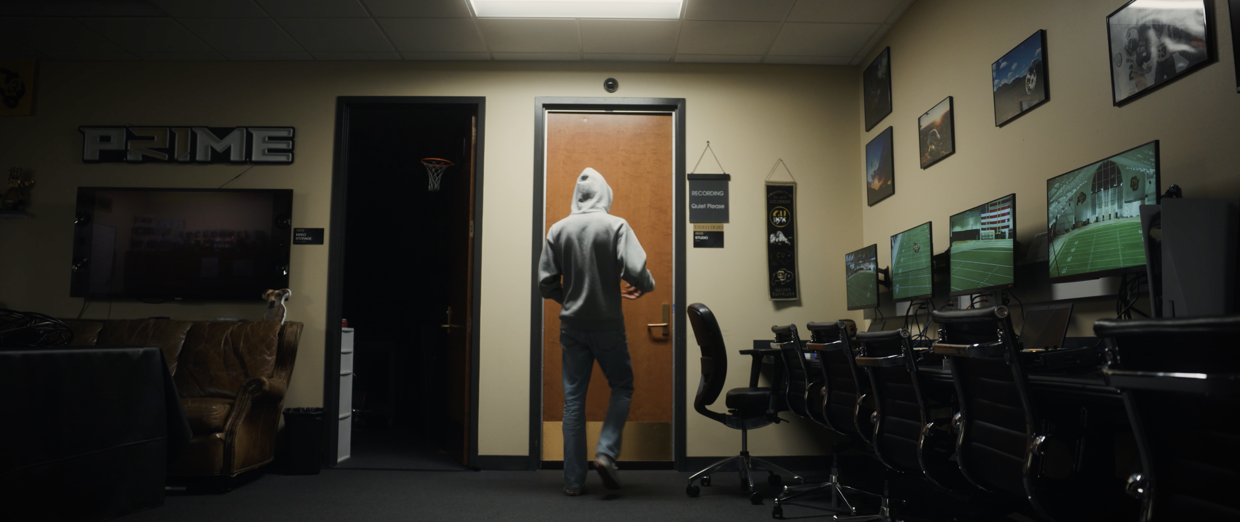 A person wearing a gray hoodie and jeans walking into a room with multiple computer monitors displaying football games, a leather couch with a small dog sitting on it, and framed pictures on the wall.