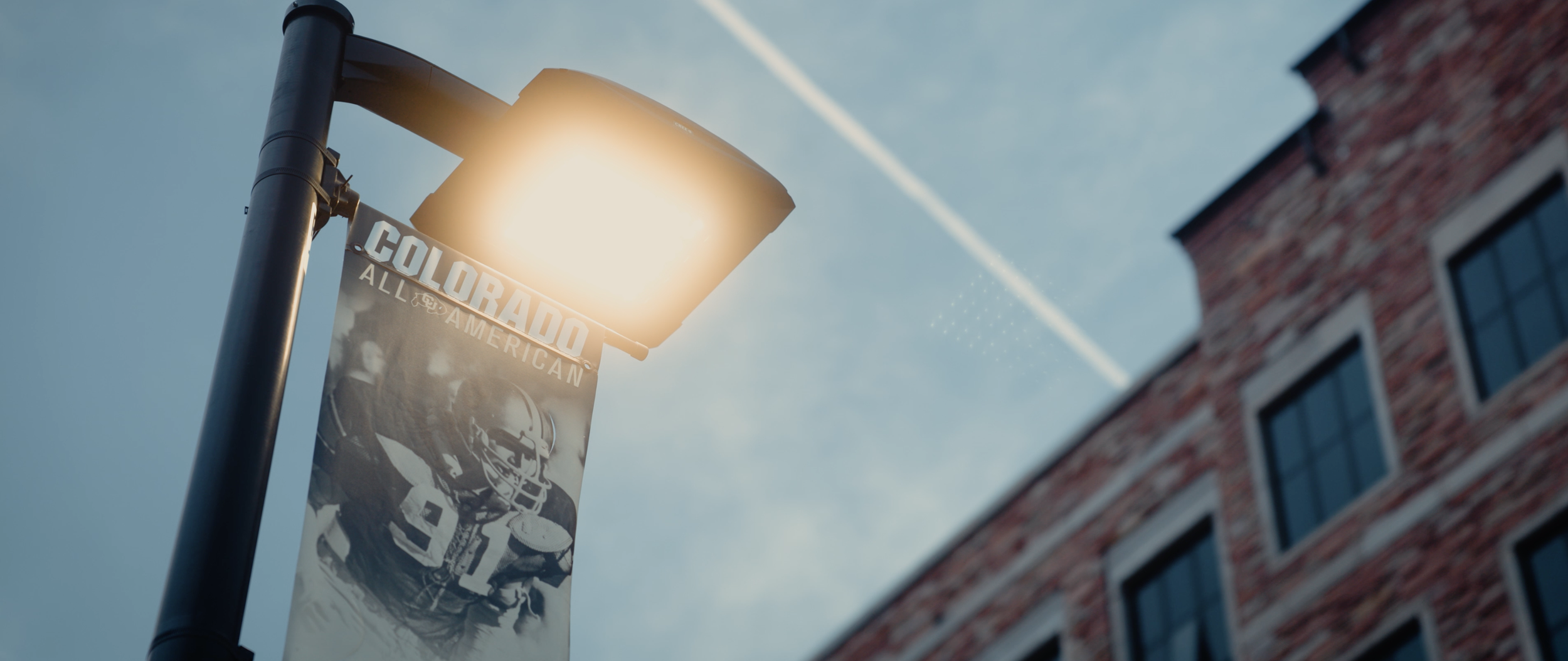 Streetlight illuminating a banner that reads "Colorado All American" with an American football player image, against a clear blue sky with a building on the right.