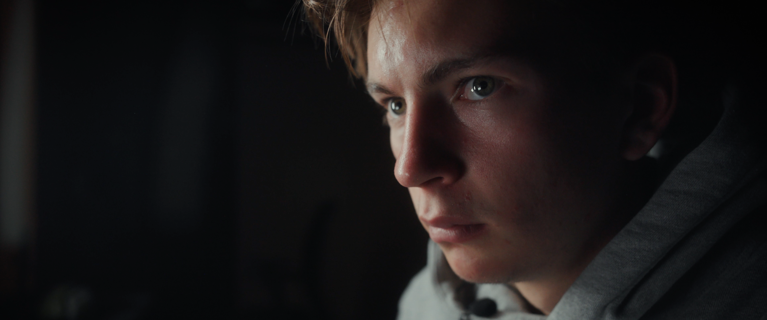 Close-up of a young man with blond hair and blue eyes, looking intently to the side in a dark setting.