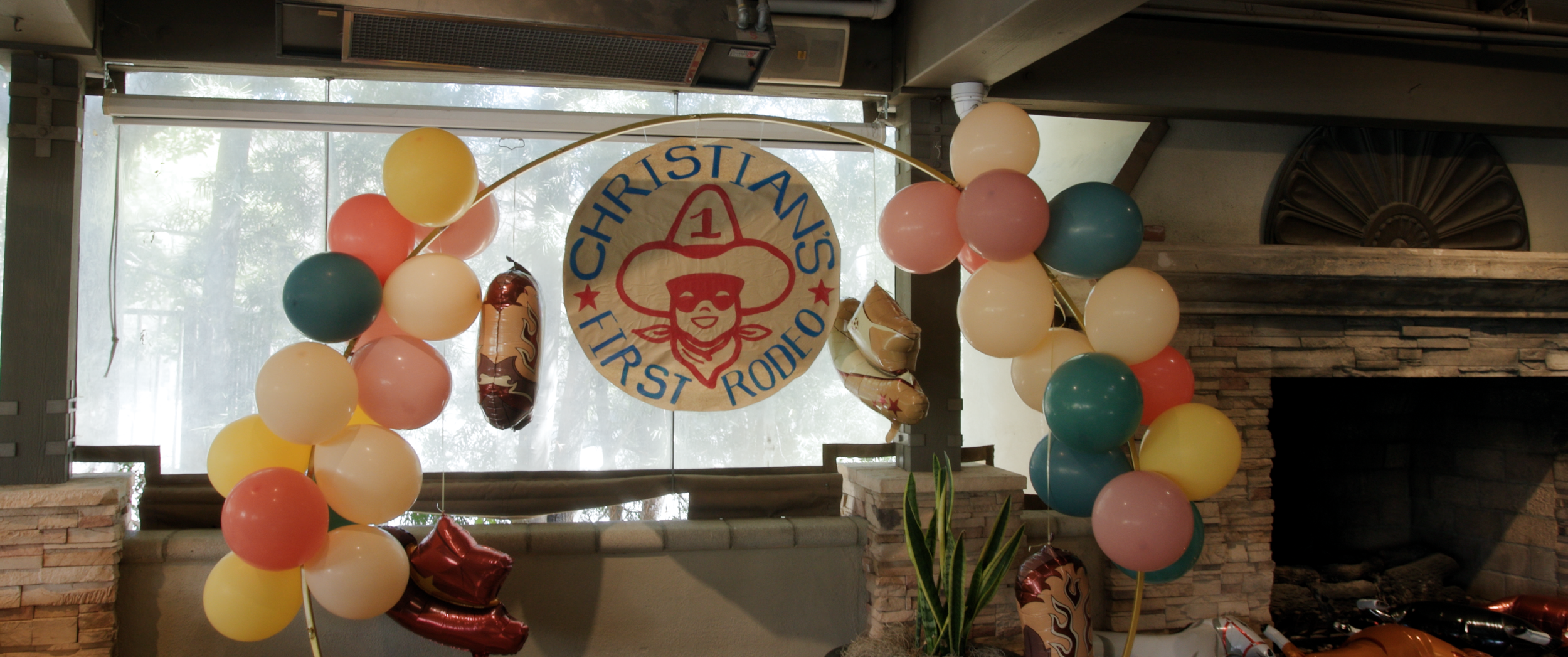 Decorations for a first rodeo celebration featuring a circular sign with a cowgirl hat and face with a bandana, the words 'Christians First Rodeo,' colorful balloons in pink, yellow, blue, and white, and foil balloons resembling cowboy boots and a wh