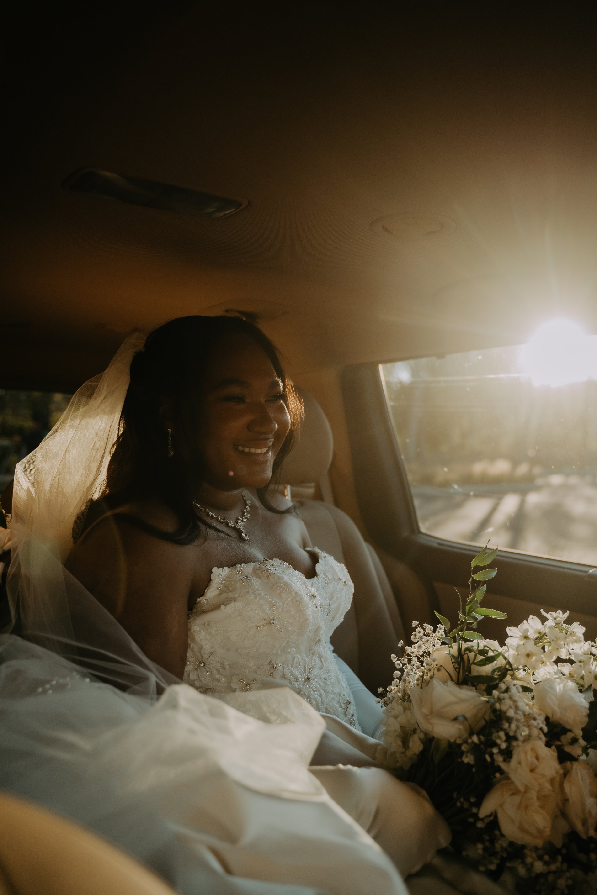 A bride smiling inside a vehicle, illuminated by sunlight, holding a bouquet of white flowers.