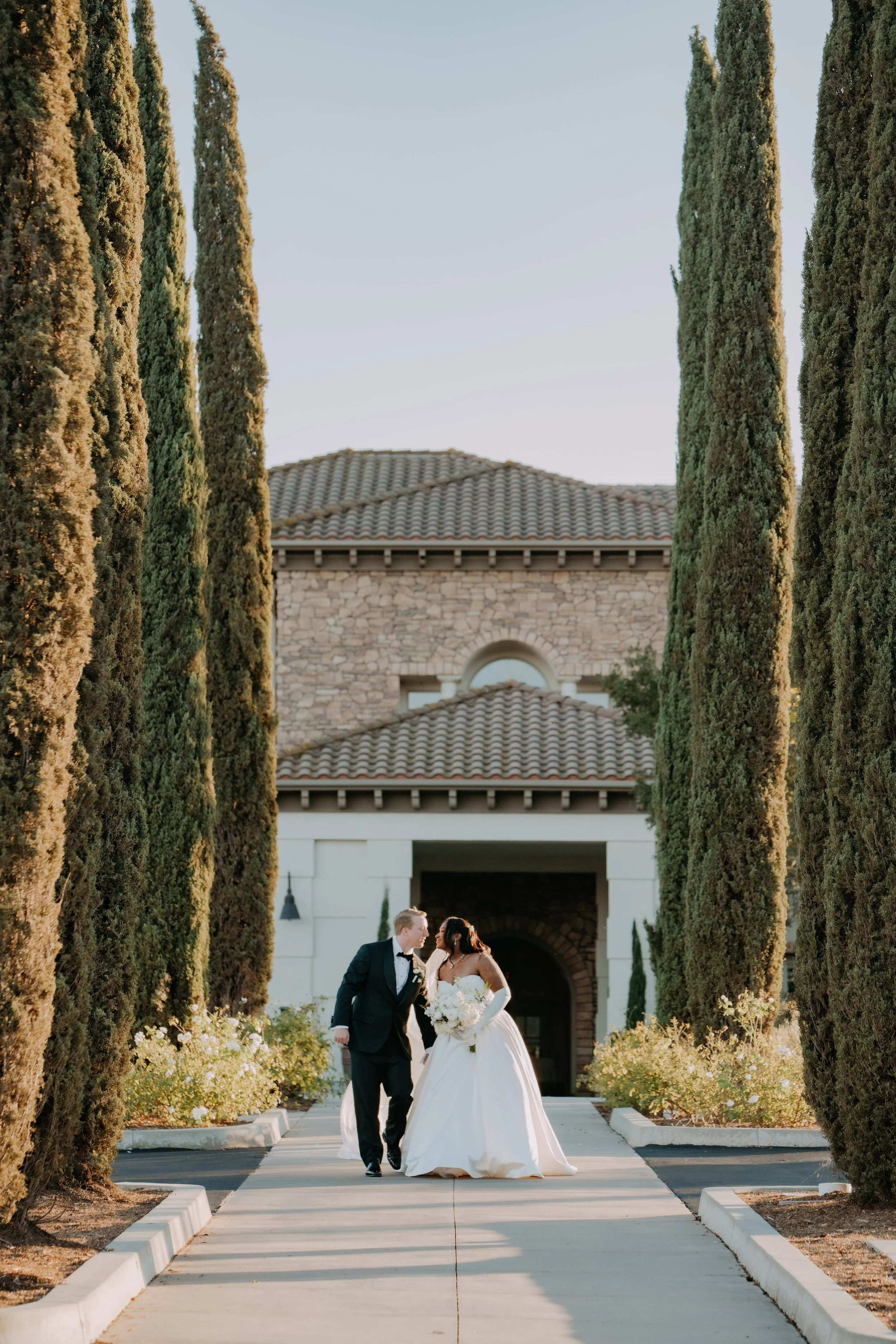 A bride and groom walk arm-in-arm down a path lined with tall cypress trees, with a large stone and stucco building in the background, during a wedding ceremony or photo session.