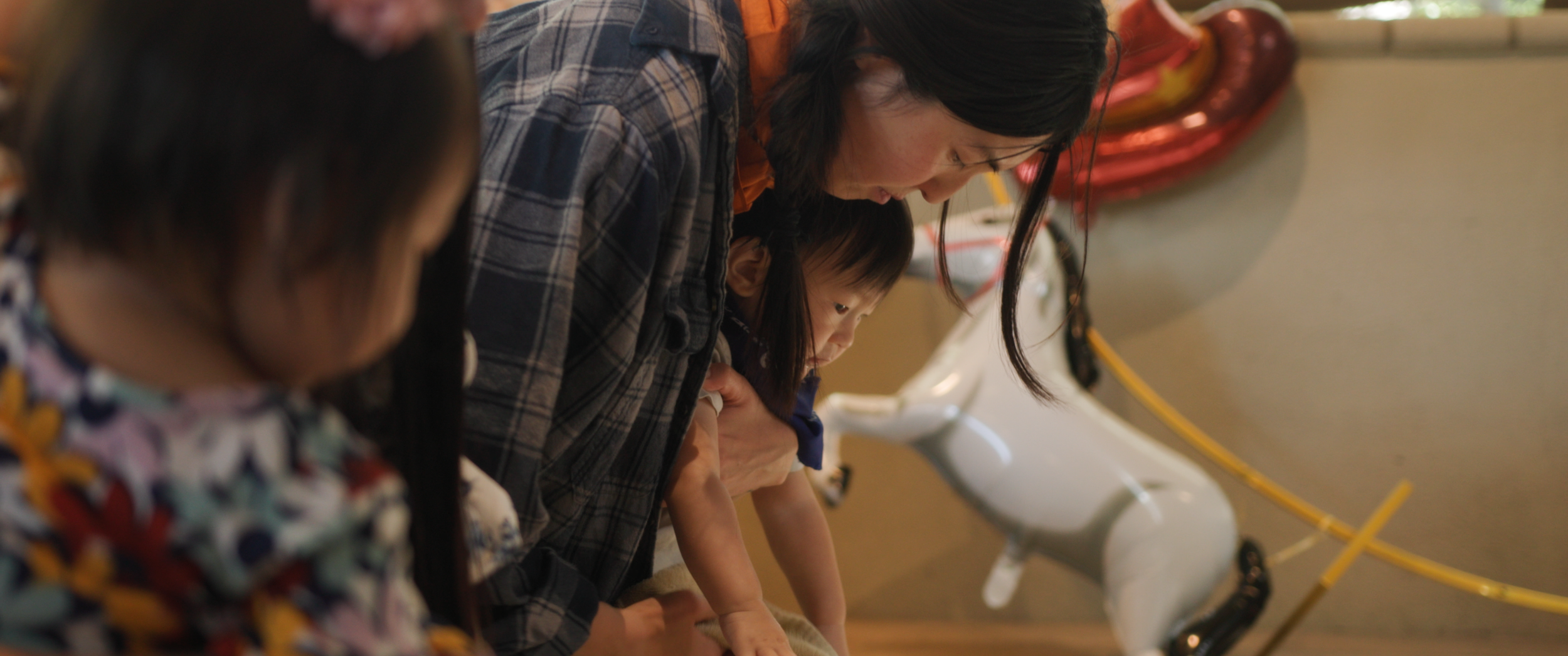 A woman and two children lying on the floor, looking at something intently, with a partial view of another child in the foreground. The background includes a wall, a model airplane, and some wires.