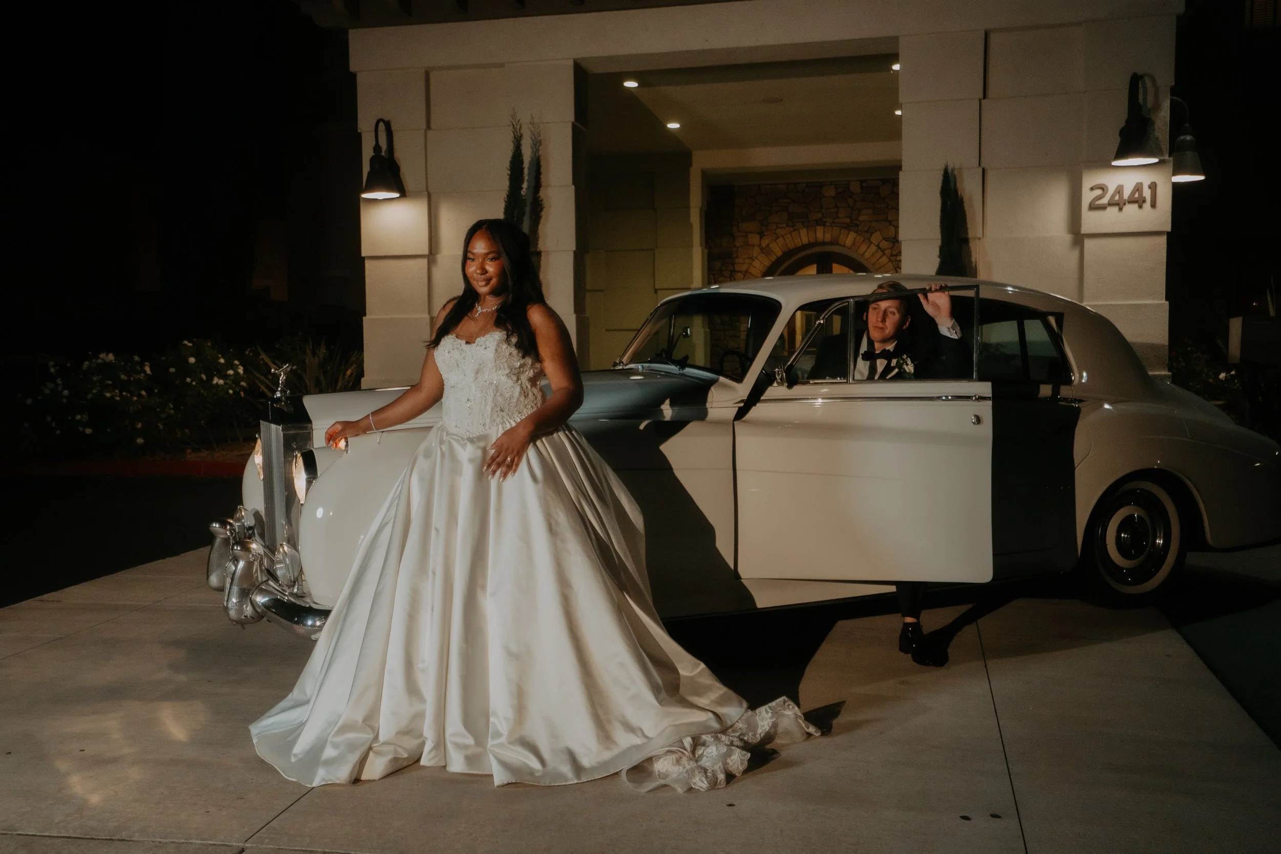Bride in wedding dress standing next to vintage white car with open door, groom sitting inside car at night in front of a building with the number 2441 visible.