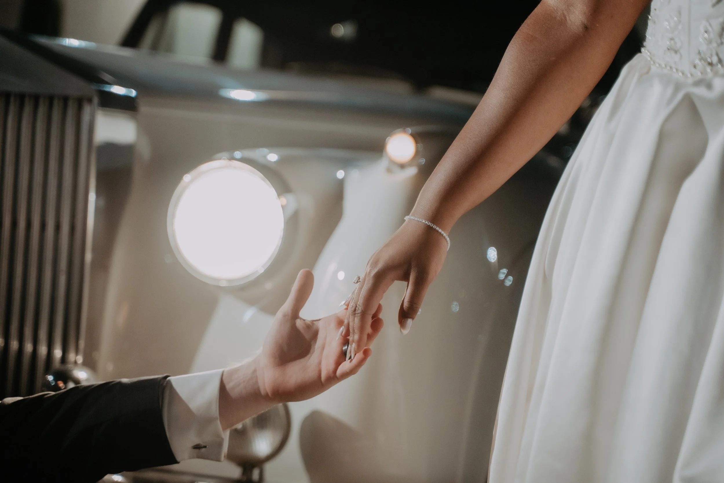 A bride touching her groom's hand during a wedding ceremony. The bride is wearing a white wedding dress and a bracelet, while the groom's suit sleeve is partially visible.