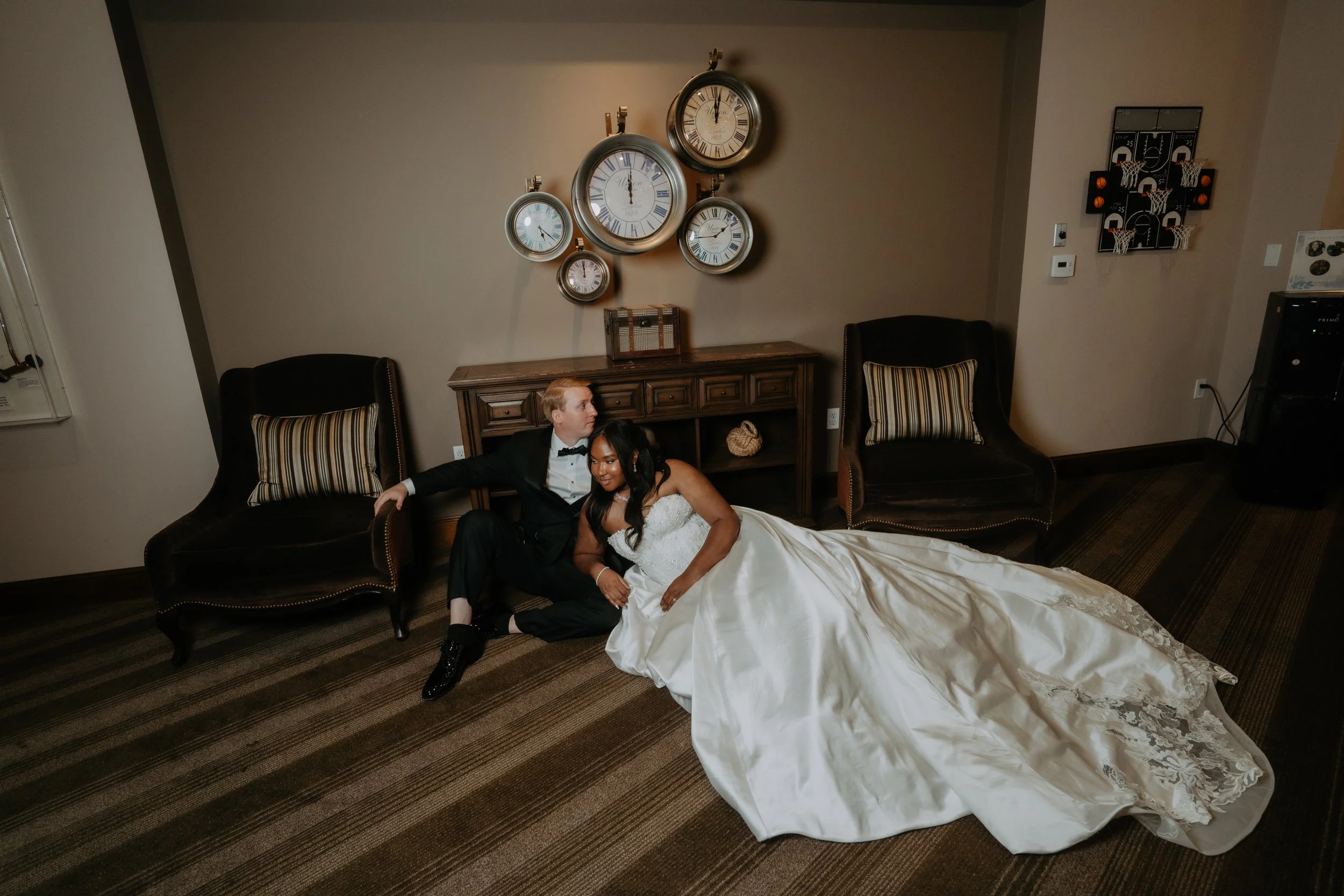 A bride and groom sitting on the floor in a room with wall clocks, two armchairs, and a wooden table, during their wedding celebration.