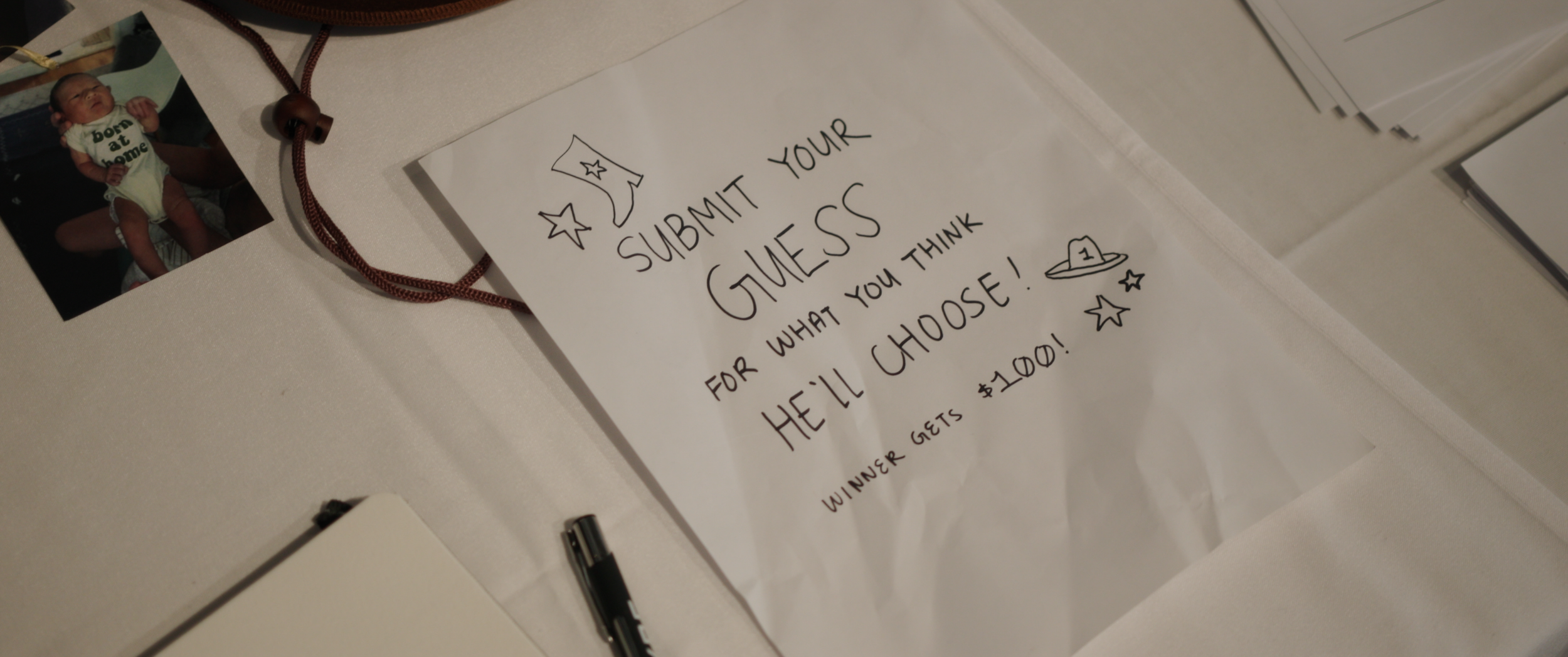 A white sheet of paper with handwritten text and drawings, placed on a table. The paper encourages submitting guesses for a contest with illustrations of a moon, stars, a hat, and a UFO. Next to the paper is a photograph of a newborn baby wearing a o