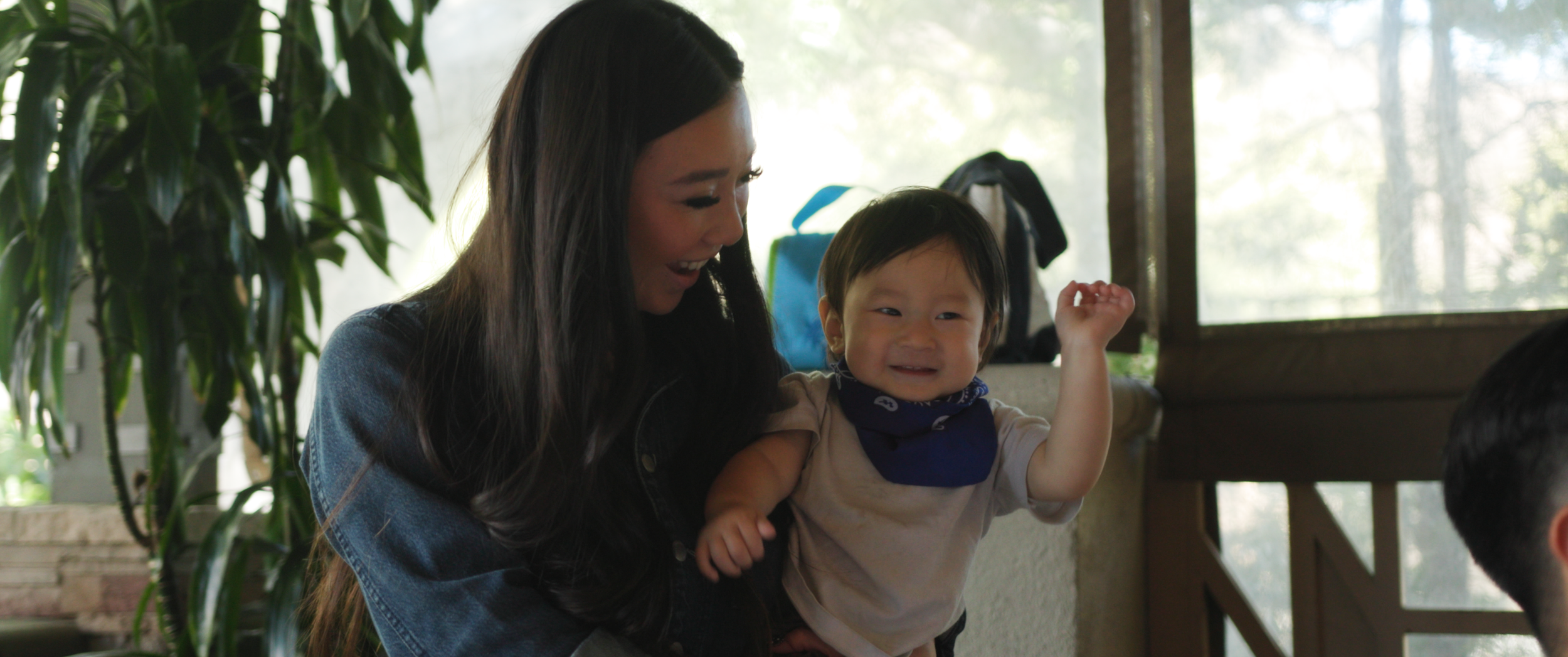 A woman holding a smiling young boy inside a room with large windows and greenery outside, with her looking at him affectionately.