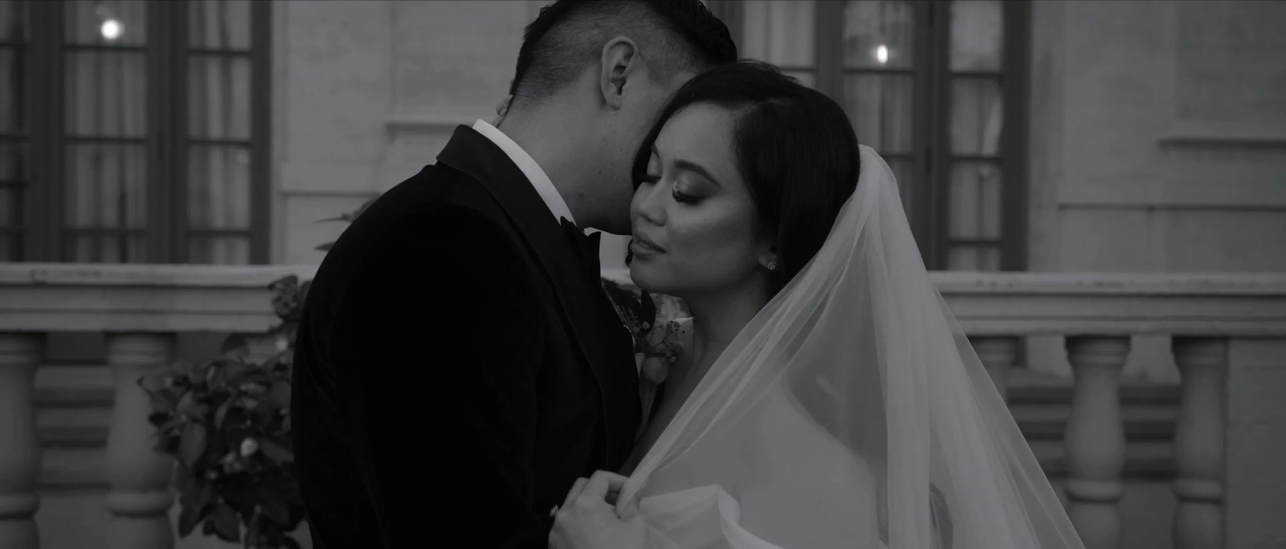 Black and white photo of a groom embracing a bride, both with closed eyes, against a building backdrop.
