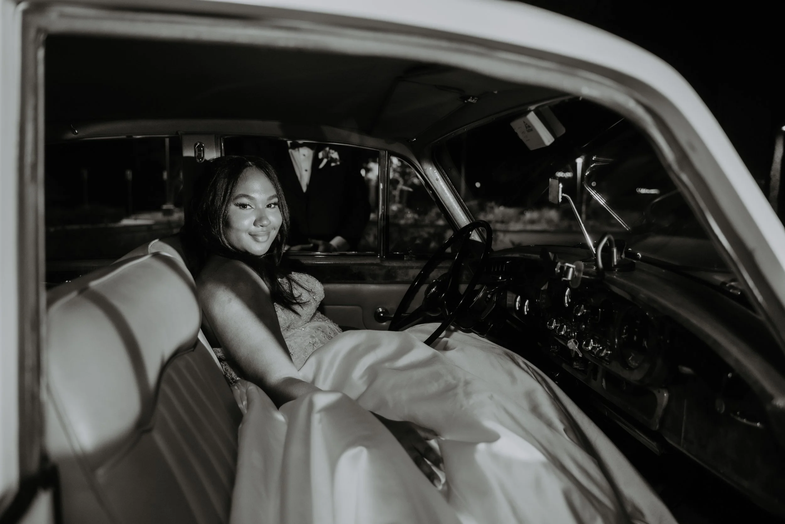 A woman in a wedding dress sitting inside a vintage car at night, smiling at the camera, with a man in a tuxedo standing outside the car in the background.