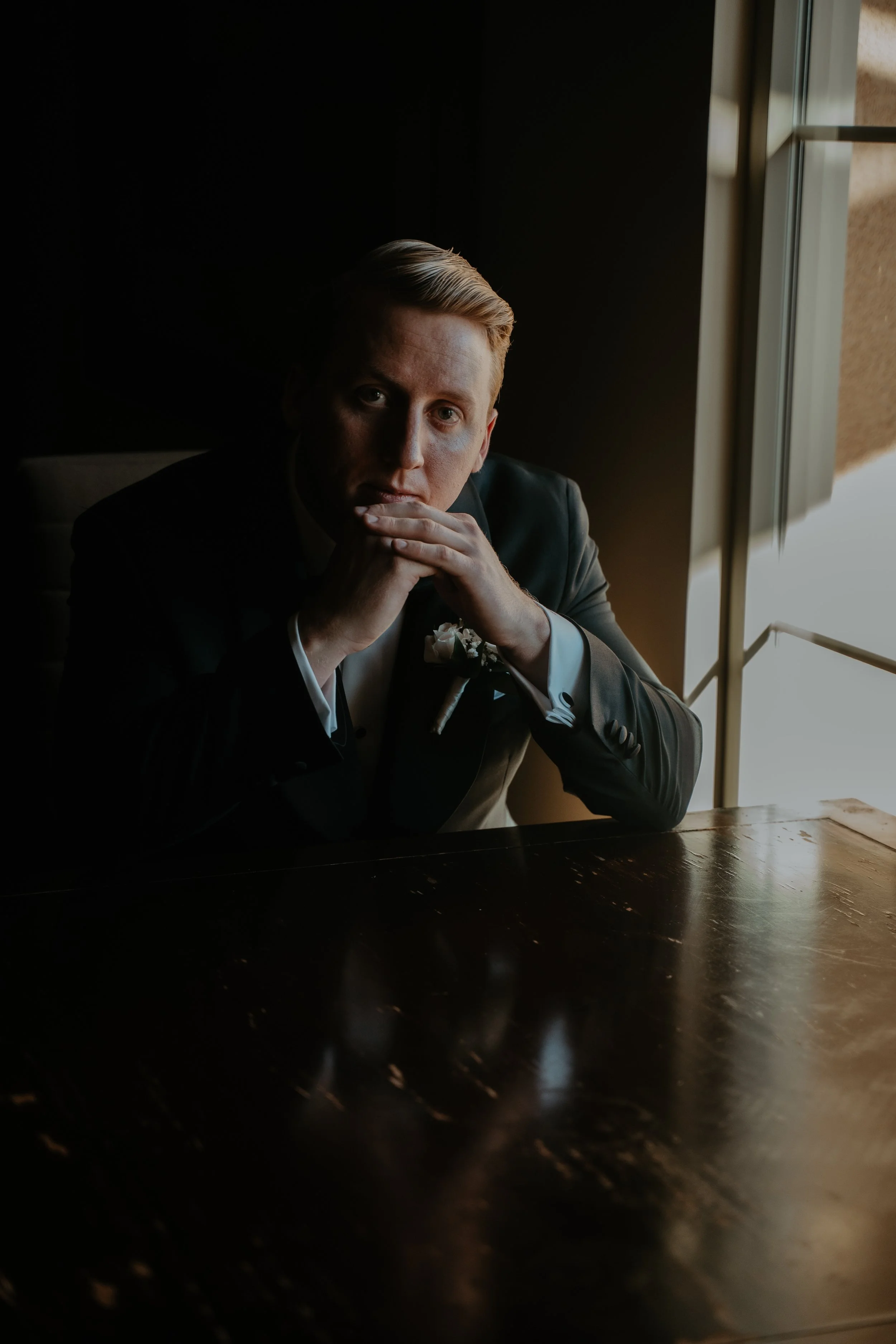 A man with blond hair sitting at a dark wooden table near a window, wearing a tuxedo, with his hands clasped near his face, looking into the camera.
